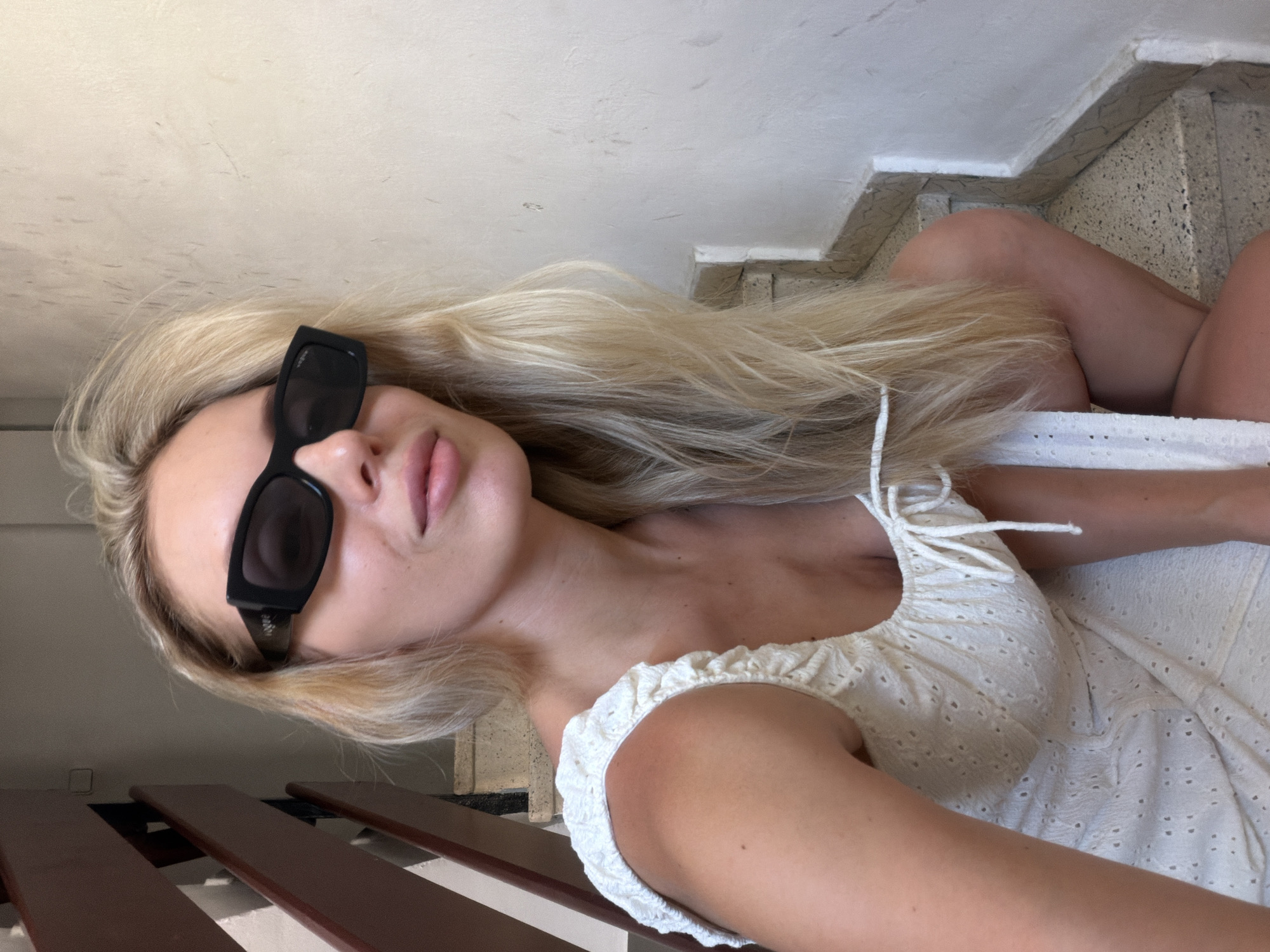 The media file shows a young woman with long blonde hair and sunglasses, wearing a white eyelet dress. She is lying on a set of wooden benches that appear to be part of a staircase. The staircase itself has speckled stone steps and white trim. The lighting suggests it is daytime, possibly outdoors. The woman's pose is relaxed, and her expression is neutral. The image is taken from a high angle, looking down at the subject. The overall impression is casual and summery.