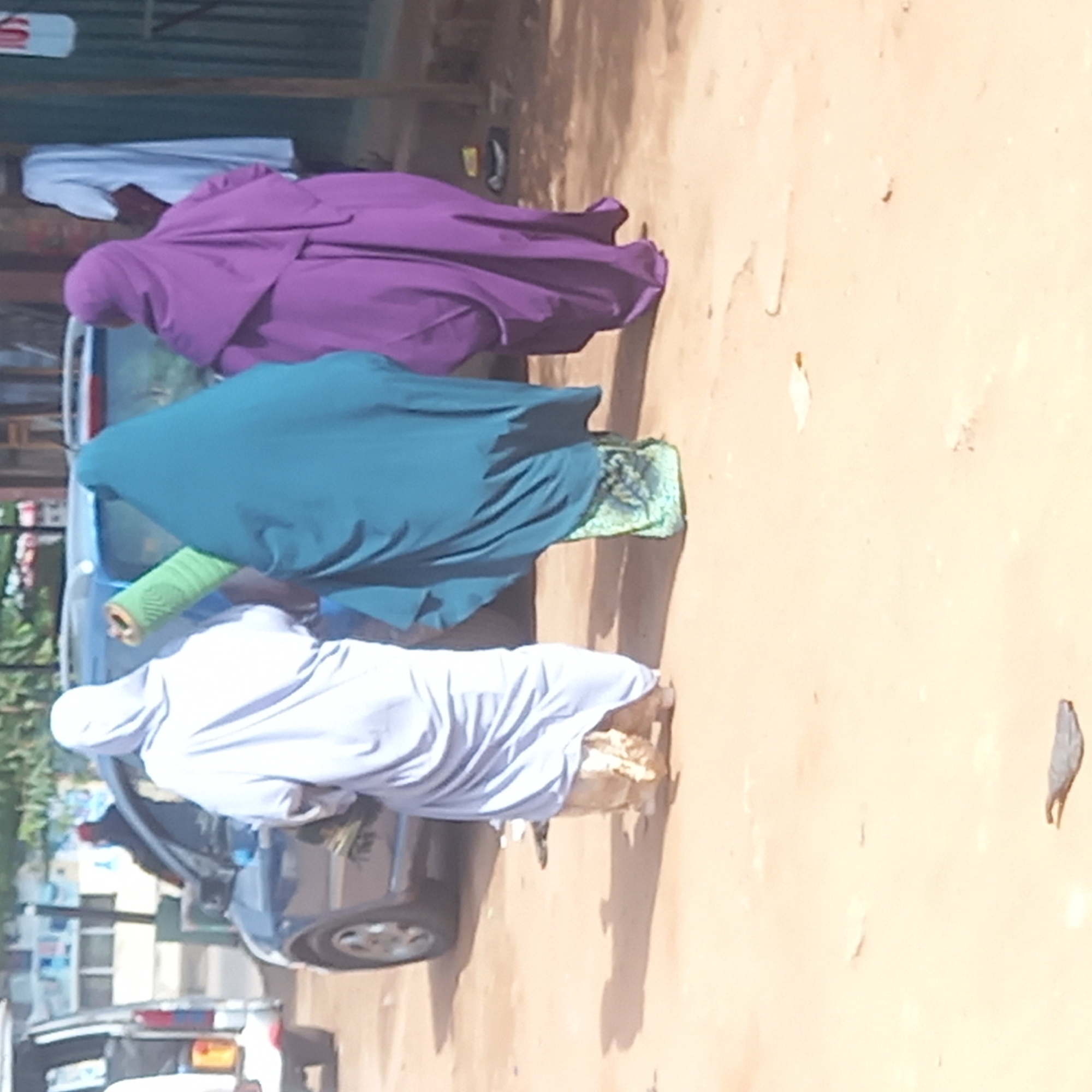 The image shows three women wearing Islamic full-body veils ("hijabs") of different colors (purple, teal, and white) walking on a dirt road in Dadi Riba, Nigeria. They are moving away from a parked grey vehicle. Other vehicles and buildings can be seen in the background.