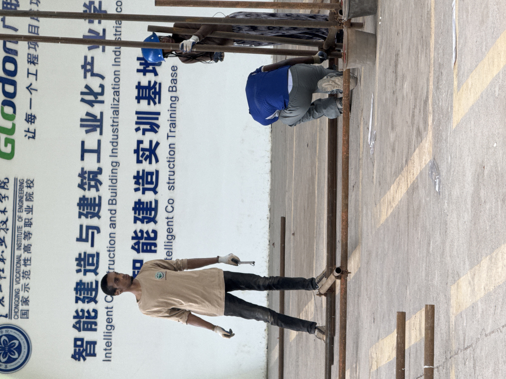 The image depicts an outdoor scene at the Chongqing Vocational Institute of Engineering in Jiangjin, China. In the foreground, two individuals appear to be engaged in construction or maintenance work. One person, wearing a light brown long-sleeved shirt and dark pants, stands with a tool in their hand, looking to the right. Another person, wearing a blue shirt and gray pants, is kneeling and working with metal pipes. A third person, wearing a dark top and patterned skirt, stands behind them, also seemingly involved in the activity. The background features a large white banner with Chinese and English text. The ground is a concrete surface with yellow markings. Several metal pipes are laid out on the ground, suggesting ongoing construction or assembly. Two grey buckets are visible near the standing figure on the right.