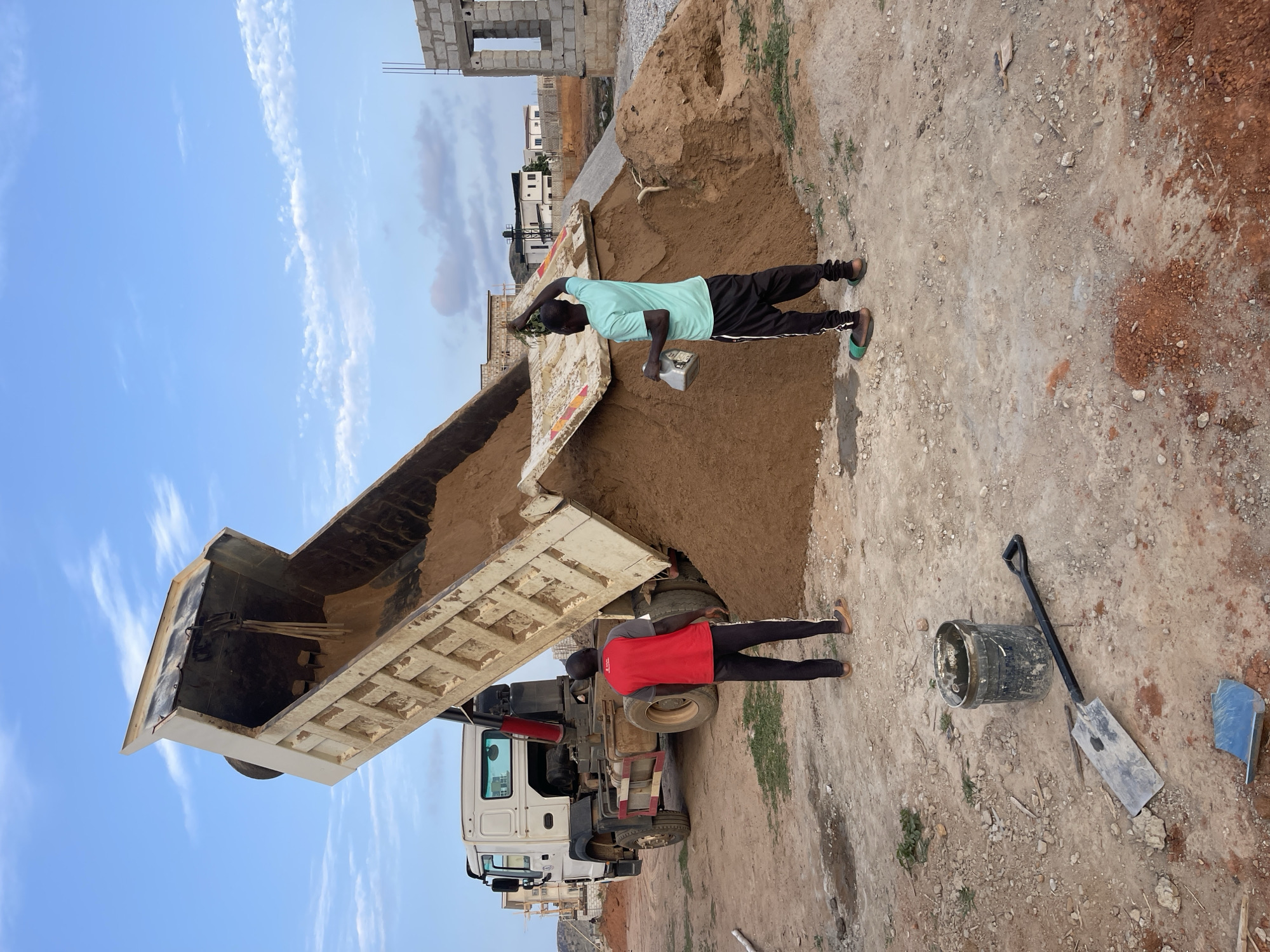 The image depicts a construction site in Angwan Sayawa, Nigeria. A large, white dump truck with its bed raised is unloading a pile of sand onto a dirt surface. Two men are present. One man wearing a light blue shirt and dark pants stands near the sand pile. The second man, wearing a red shirt and dark pants, stands near the truck's rear tires. A bucket, shovel, and trowel are on the ground near the truck. In the background, a partially constructed building is visible, as well as other buildings and a blue sky with scattered clouds. The ground surface is mostly dirt and gravel.