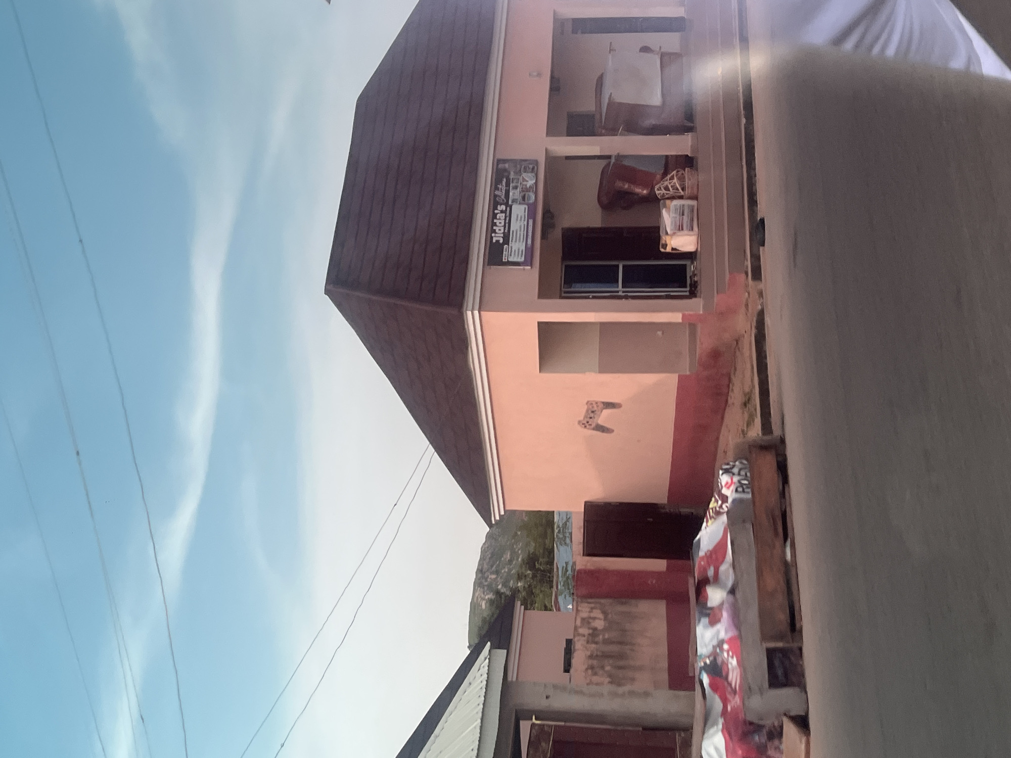This is an outdoor, eye-level, rotated image of a pink building in Jalingo, Nigeria. The building has a brown, sloped roof and a sign that reads "Jidda's Collection". The scene appears to be during the daytime, with a light blue sky and some wispy clouds. There are electrical wires visible in the sky. The building has several windows and doors, and there are some items stored outside, including what looks like a basket and some bundled items. The overall impression is of a shop or business in a tropical climate.
