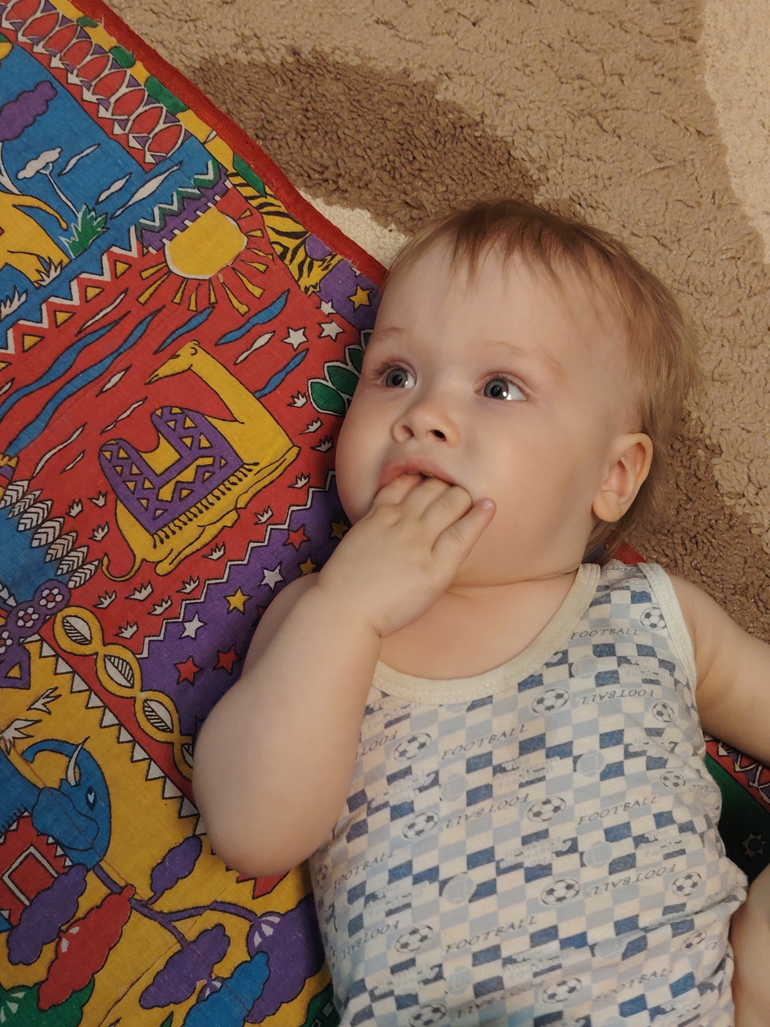 A baby lies on a colorful rug and a beige carpet. The rug features stylized animal and sun designs in blue, red, yellow, and purple. The baby has fair skin, blonde hair, and is wearing a light blue and white checkered tank top with a football print. The baby is looking upwards and has its hand near its mouth.
There is no information to confirm the location of this image is in Chaikovsky, Russia.