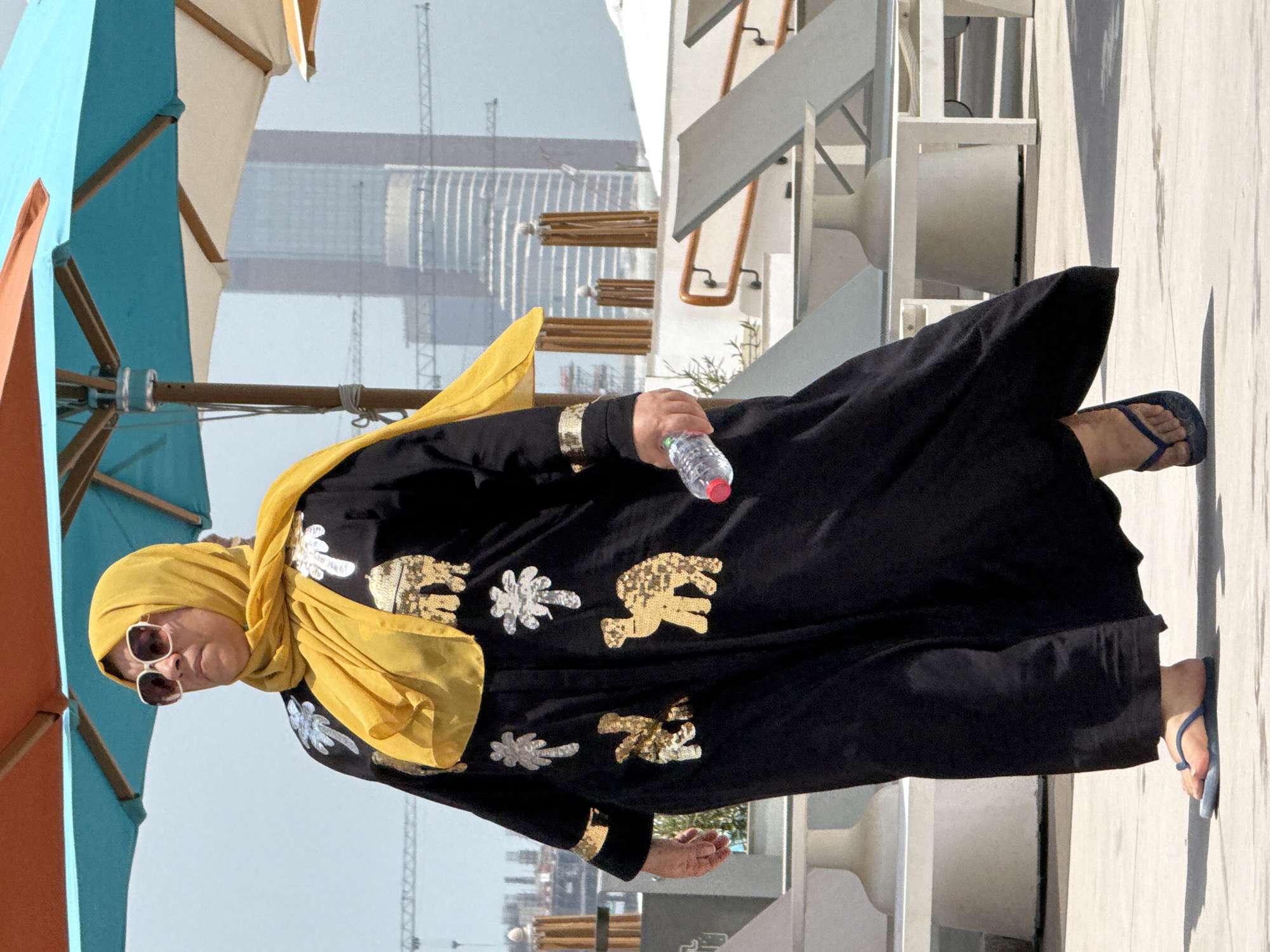 An adult woman wearing a yellow hijab and a black abaya adorned with gold sequined camel and palm tree motifs walks outdoors. She is wearing large sunglasses and blue flip-flops. In her right hand, she holds a clear plastic bottle of water with a red cap. The woman is walking on a paved surface, with lounge chairs and umbrellas visible in the background. In the distance, a tall skyscraper and construction cranes are visible, suggesting an urban setting in the United Arab Emirates. The lighting indicates it is daytime.