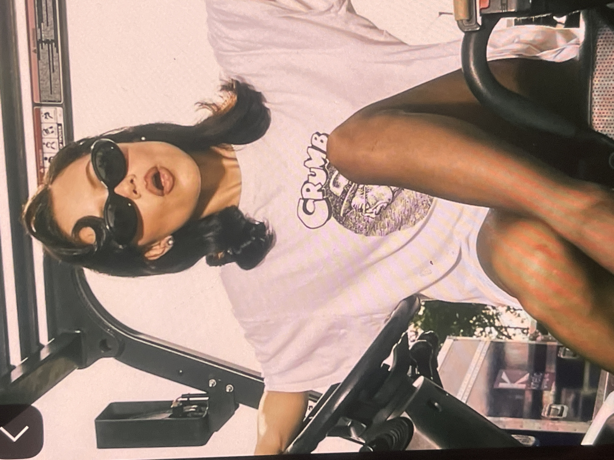 The image, rotated 90 degrees counter-clockwise, features a young woman posing against a backdrop of what appears to be equipment and an outdoor scene.
**Subjects:** The central subject is a woman with dark hair, visible from the waist up. She is wearing large, black, oval-shaped sunglasses and a white, short-sleeved t-shirt. Her mouth is slightly open, and she looks directly towards the viewer.
**Objects:** Her white t-shirt prominently displays the word "CRUMB" in a distinctive, stylized font, accompanied by a circular graphic underneath featuring a dark, cartoonish illustration, possibly of a character or creature. The woman is positioned amidst dark, metallic structures, likely part of gym equipment, industrial machinery, or a vehicle interior, such as a large van or RV. A black handlebar or steering wheel-like component is visible near her right arm (viewer's left), and a small, dark tray or compartment is in the lower left foreground.
**Scene/Setting:** The immediate setting is an interior or semi-enclosed space, defined by the dark equipment. Beyond this, through an opening or window, an outdoor environment is visible. This includes green foliage from trees and parts of a light-colored structure or vehicle, possibly with visible shelving or internal details. The overall impression is casual and somewhat urban or semi-rural, given the trees.
**Activity or event taking place:** The woman is captured in a posed moment, looking directly at the camera with a playful or expressive facial gesture. She is not actively engaged in using the equipment but rather reclining or leaning on it for the photograph.
**Notable details:** The photograph appears to be taken during the daytime, suggested by the natural light illuminating the background. The woman's expression, combined with her sunglasses, conveys a cool and confident demeanor. The image itself has a somewhat soft or slightly grainy aesthetic. While the location is provided as Poltava, Ukraine, there are no specific visual cues within the image to explicitly identify this city.
**Visible text:** The word "CRUMB" is clearly visible on the front of the woman's white t-shirt. No other readable text is present in the image.