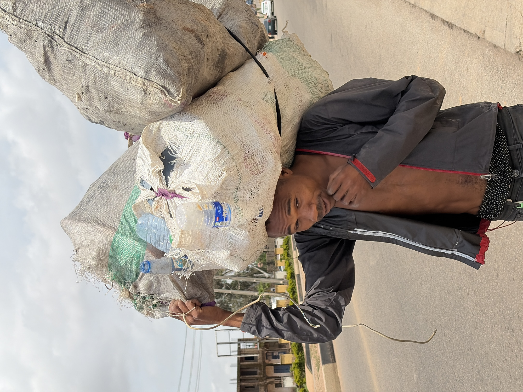 A man is carrying a large load of sacks on his head and shoulders, walking along a paved road. He is wearing a dark jacket over a bare chest and dark pants with a patterned waistband. The sacks appear to be made of rough material and contain various items, including plastic bottles. The man's face is visible, and he appears to be looking forward.
The scene is outdoors, with a road and some buildings in the background. The sky is cloudy. There is a slight overcast, suggesting it could be daytime, possibly morning or afternoon. The location is identified as Gura Top, Nigeria.
The overall impression is one of hard labor and the carrying of goods, likely for sale or transport. The man's expression is neutral, with no strong indication of joy or distress. The activity appears to be mundane, a daily task for the individual. The presence of plastic bottles suggests the possibility of collecting recyclables or transporting goods in plastic containers.