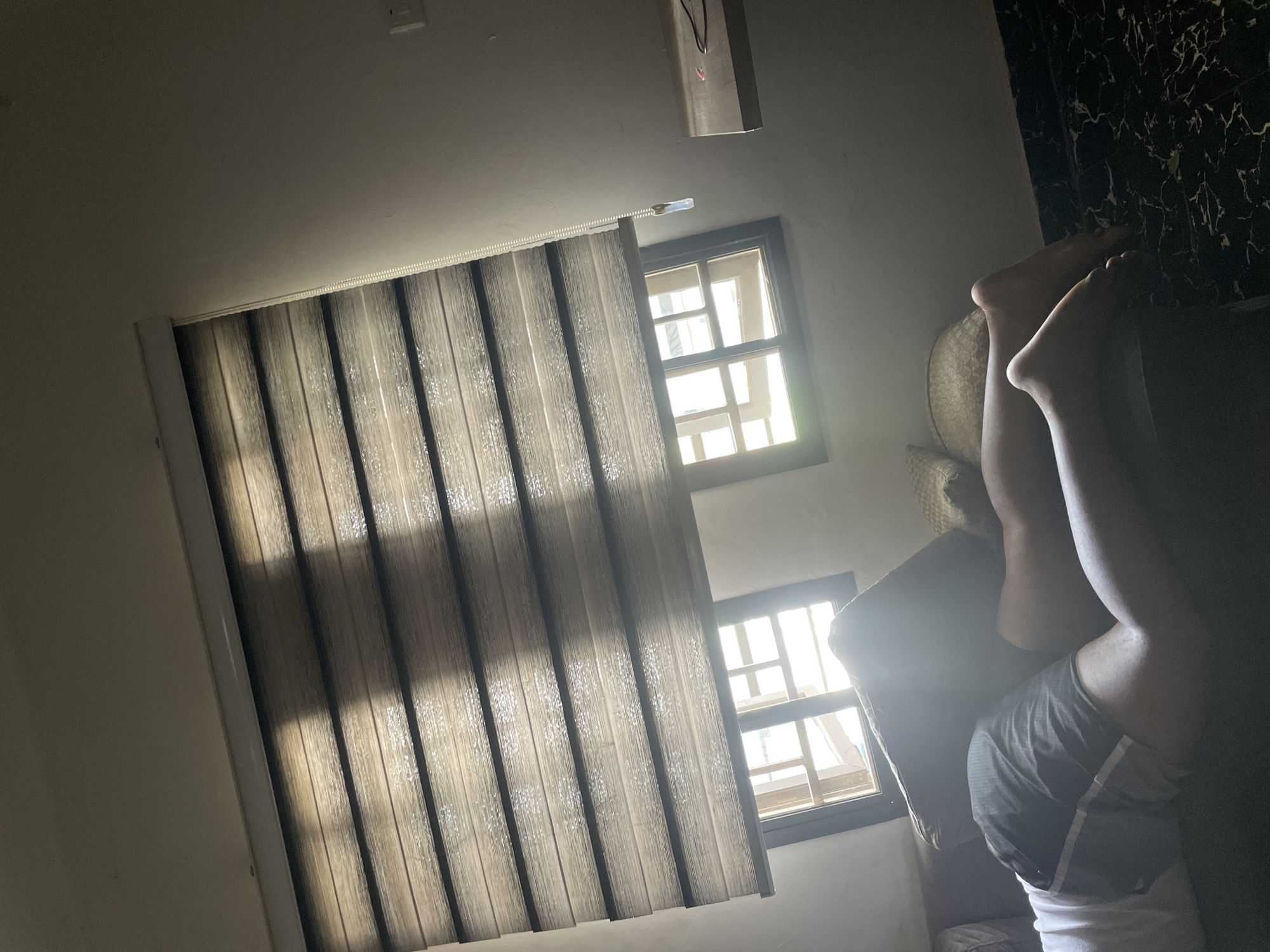 The interior of a room in Abuja, Nigeria, is seen. A vertical blind partially covers a window, revealing sunlight. A second window is visible next to the blinds. A person is lying on a sofa, with their legs elevated. They are wearing shorts and a white shirt. A hat rests on the sofa next to the person. The background is a patterned wall.