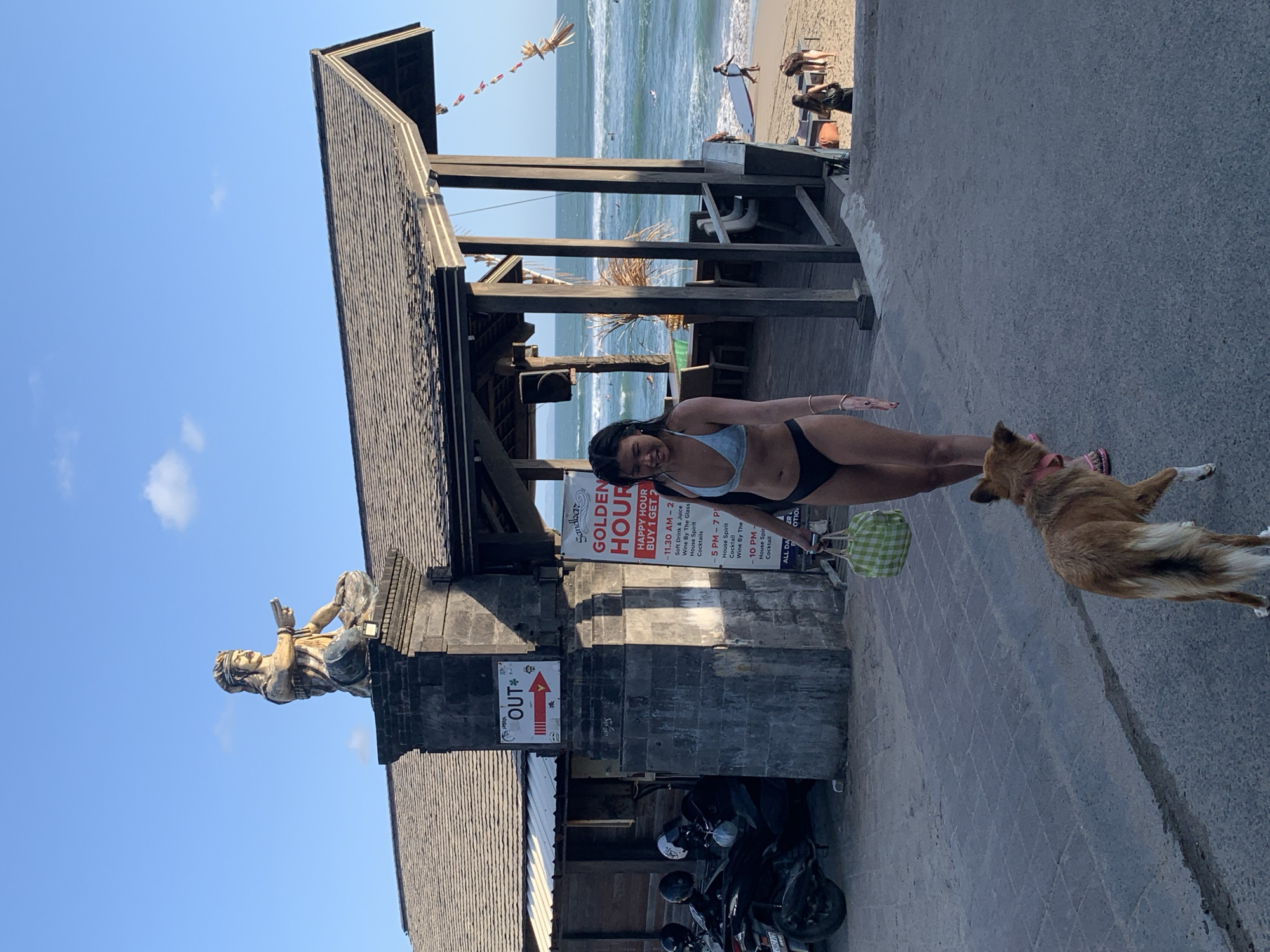 A woman in a bikini walks on a paved path towards the viewer, holding a green checkered bag. A medium-sized brown dog with a pink collar walks beside her. In the background, a wooden structure with a thatched roof overlooks a beach with people and surfboards. A statue of a figure in a seated position is visible on top of a stone pillar to the left of the woman. A sign with red lettering indicating "GOLDEN HOUR HAPPY HOUR BUY 1 GET 2" is attached to the wooden structure. The scene appears to be a beach resort or restaurant area in North Kuta, Indonesia, under a clear blue sky with scattered clouds.
