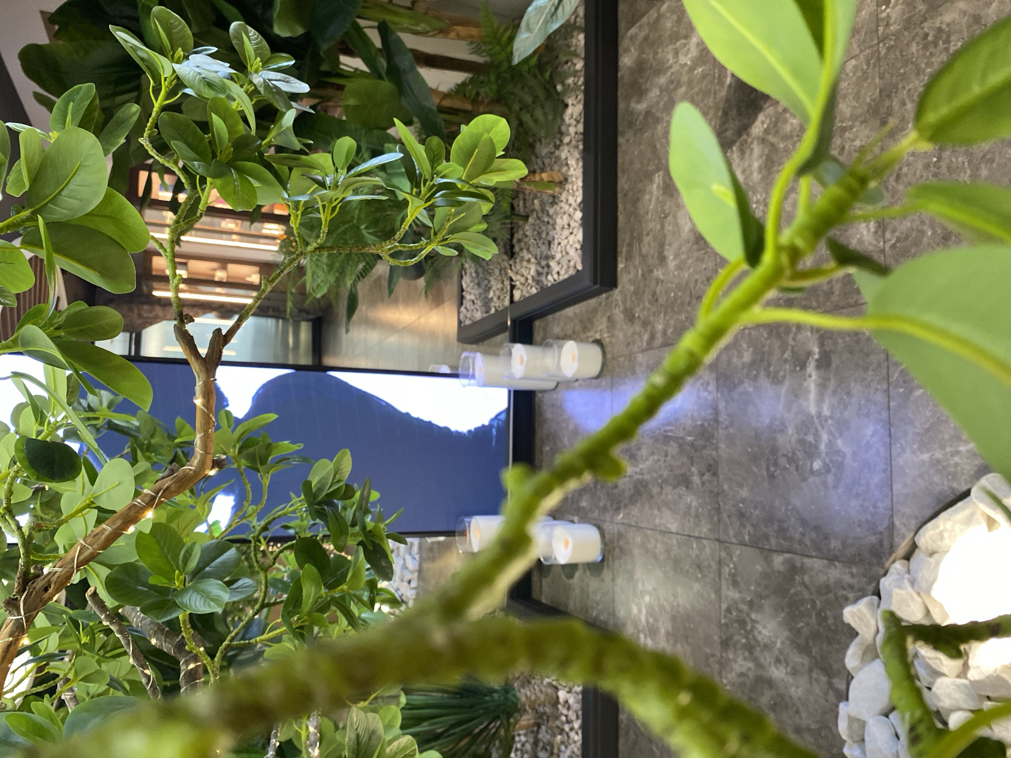 This image, taken with a slightly Dutch tilt, captures a detailed view of an indoor decorative setting. The foreground is dominated by artificial green plants with numerous leaves and branches, some of which are wrapped with delicate strings of small, warm lights.
In the mid-ground, several white, cylindrical objects resembling flameless LED candles with a soft, warm glow are arranged horizontally on what appears to be a dark, tiled surface or shelf. To the right, a section filled with white pebbles or small stones is visible, framed by a dark, possibly metal, border.
The background features a large, dark screen or display showing an abstract dark shape, possibly a silhouette of a mountain or wave, against a lighter, bluish area. Behind the plants, hints of other artificial foliage and what could be wooden slats or a textured wall are visible. The flooring throughout the visible area consists of large, grey tiles with a marble-like pattern.
No people are present, and no specific activity is taking place. The scene suggests a modern and well-decorated interior space, possibly a commercial establishment like a lobby, restaurant, or retail store, designed to create a calm and inviting atmosphere with its ambient lighting and greenery. The time of day is not discernible due to the indoor setting and artificial lighting. No visible text is present in the image.