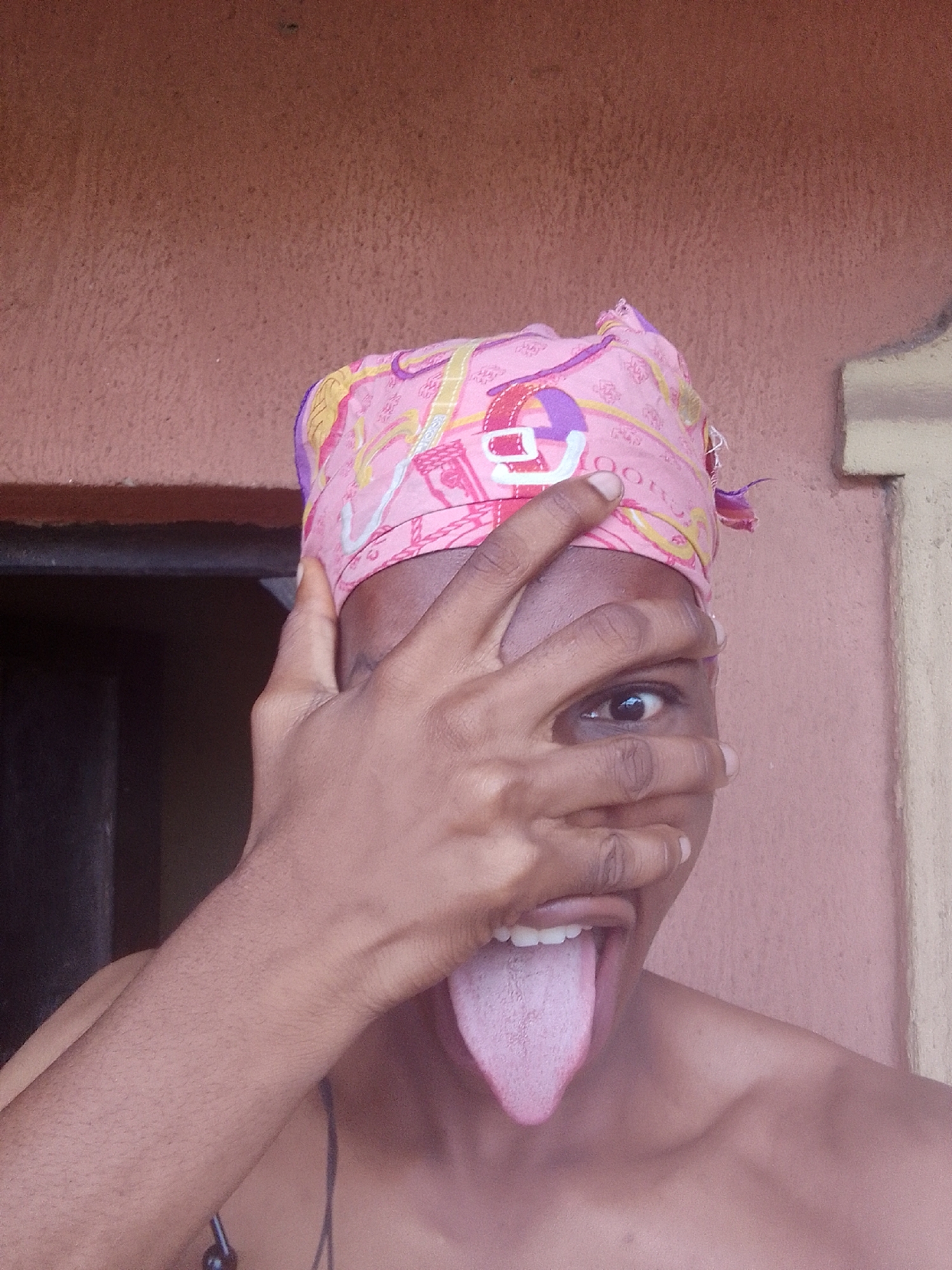 A person is standing in an outdoor location with a wall visible in the background. The person is wearing a pink head covering and has their hand partially covering their face, with one eye visible. The person's mouth is open, and their tongue is extended outward. The environment appears to be a residential area, with part of a doorway or window frame visible in the background. The specific city and location are unknown.