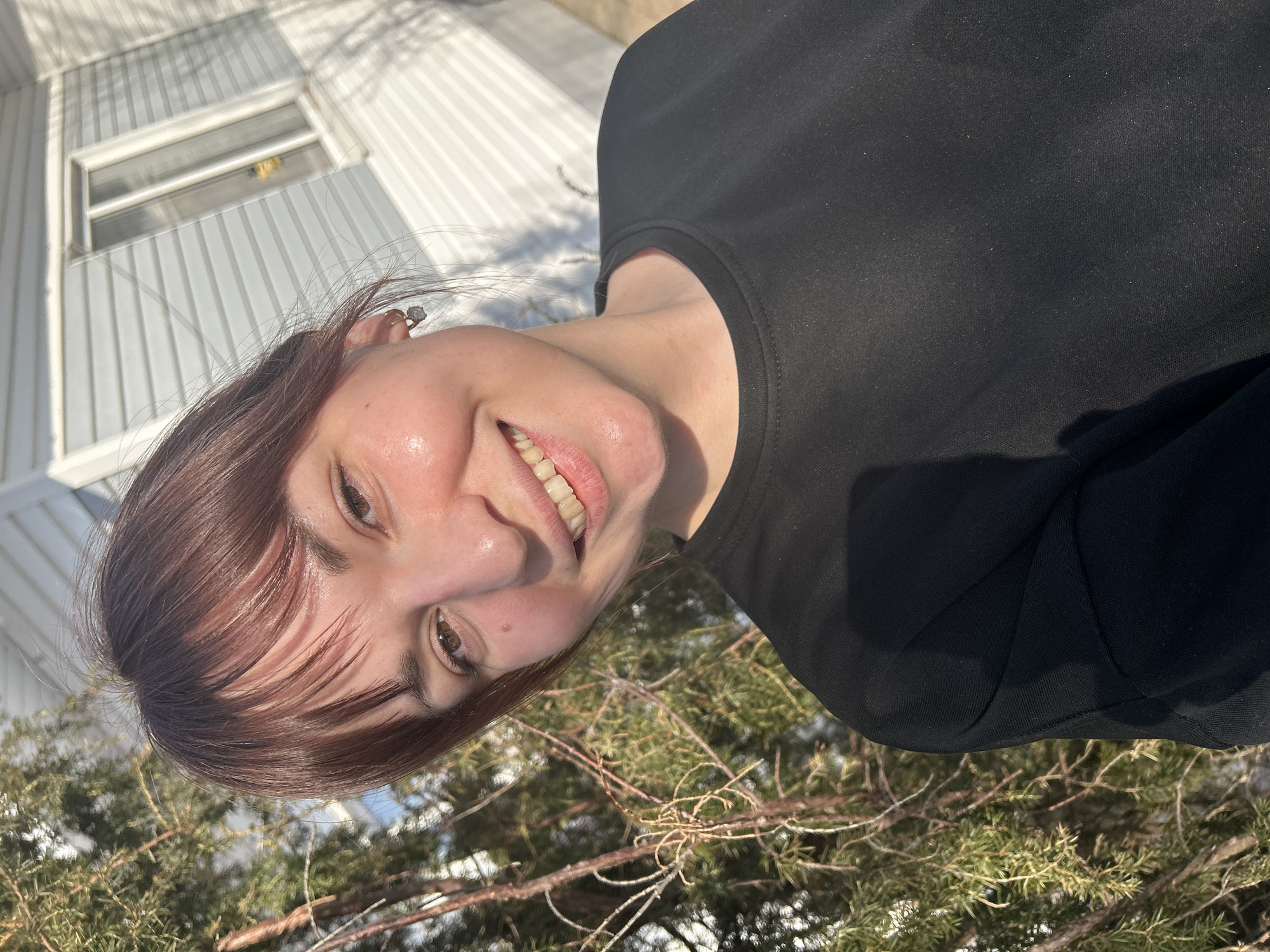 A young adult female is depicted in a close-up portrait, smiling directly at the camera. She has brown hair with bangs, brown eyes, and fair skin with a slight flush on her cheeks. She wears a plain black long-sleeved crew-neck top and small hoop earrings featuring a clear stone or charm. The background to her left features the green foliage and brown branches of a coniferous tree, suggesting an outdoor setting. To her right, a light-colored building with horizontal siding is visible, including a white-framed window. Strong sunlight is present, casting distinct shadows from tree branches onto the building. The scene is located in the city of Фоки, Russia.