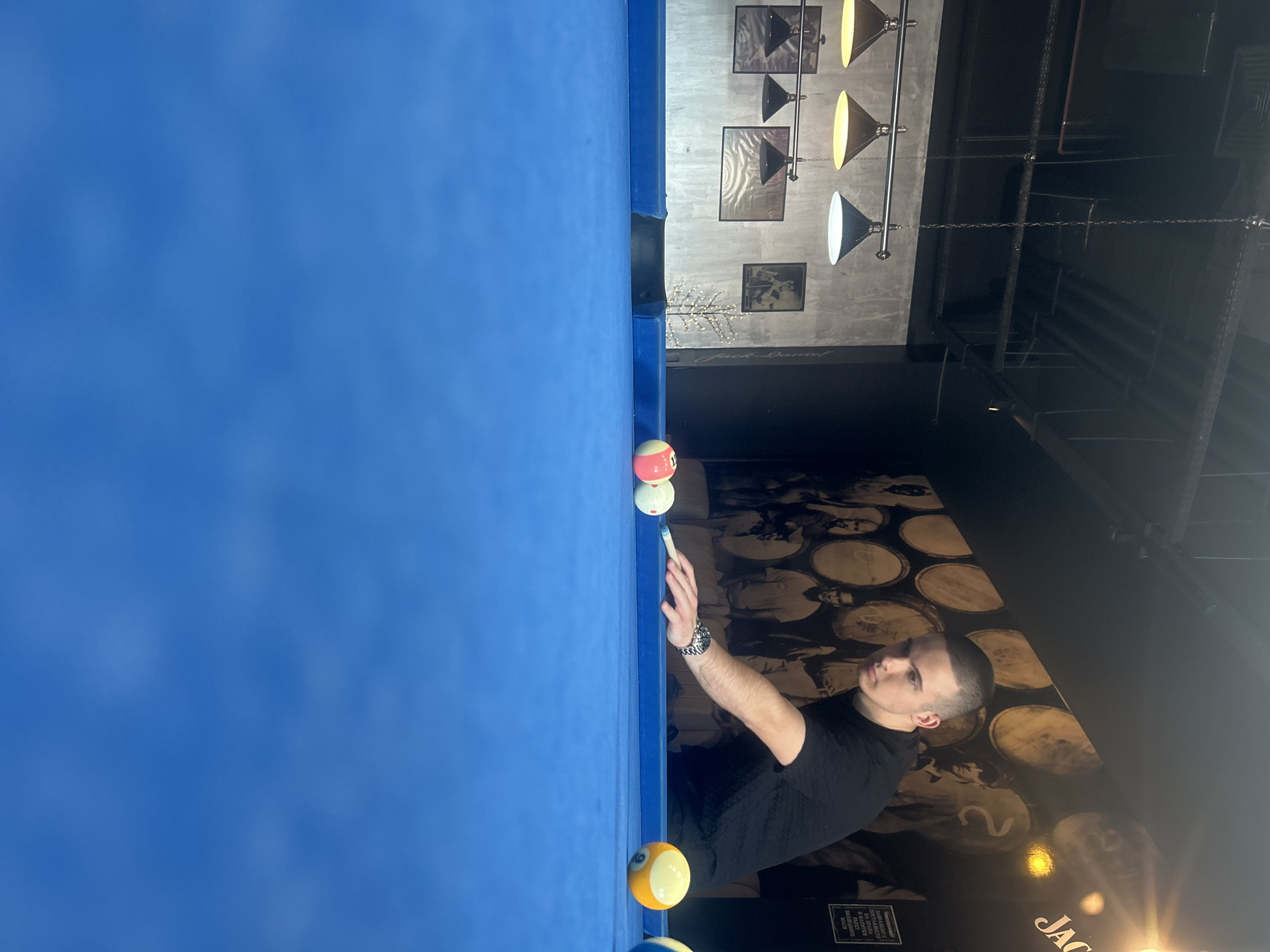 A young man is captured in a focused moment, playing pool in an indoor setting. The image is oriented vertically, showing the man leaning over a blue-felt pool table. He is wearing a black t-shirt and has short, closely cropped hair. His right arm is extended, holding a cue stick, and his left hand is forming a bridge on the table, supporting the cue. A silver watch is visible on his left wrist.
On the blue pool table, a white cue ball is positioned near his cue, ready for a shot. Other pool balls are visible, including a red and white striped 1-ball and a yellow 9-ball, both positioned closer to the viewer and the edge of the table. A pocket of the pool table can be seen further down the table from the player.
The background reveals the interior of what appears to be a bar or billiards hall. The wall behind the man features a dark mural depicting several barrel lids, some with indistinct numbers or figures. Below this mural, the brand name "Jack Daniel's" is partially visible. Further up on the wall, there are several framed pictures, and a unique light fixture resembling bare branches with small lights. Overhead, a row of industrial-style pendant lamps with conical shades provides illumination; some lights are yellow-toned, others white. The ceiling above is dark, with visible structural elements and chains supporting the lighting. A small white rectangular sign with some Cyrillic text, partially readable as "Забранено" (Forbidden), is affixed to a dark structure in the lower right background. The overall atmosphere suggests an evening of leisure. The location context is Pleven, Bulgaria.
