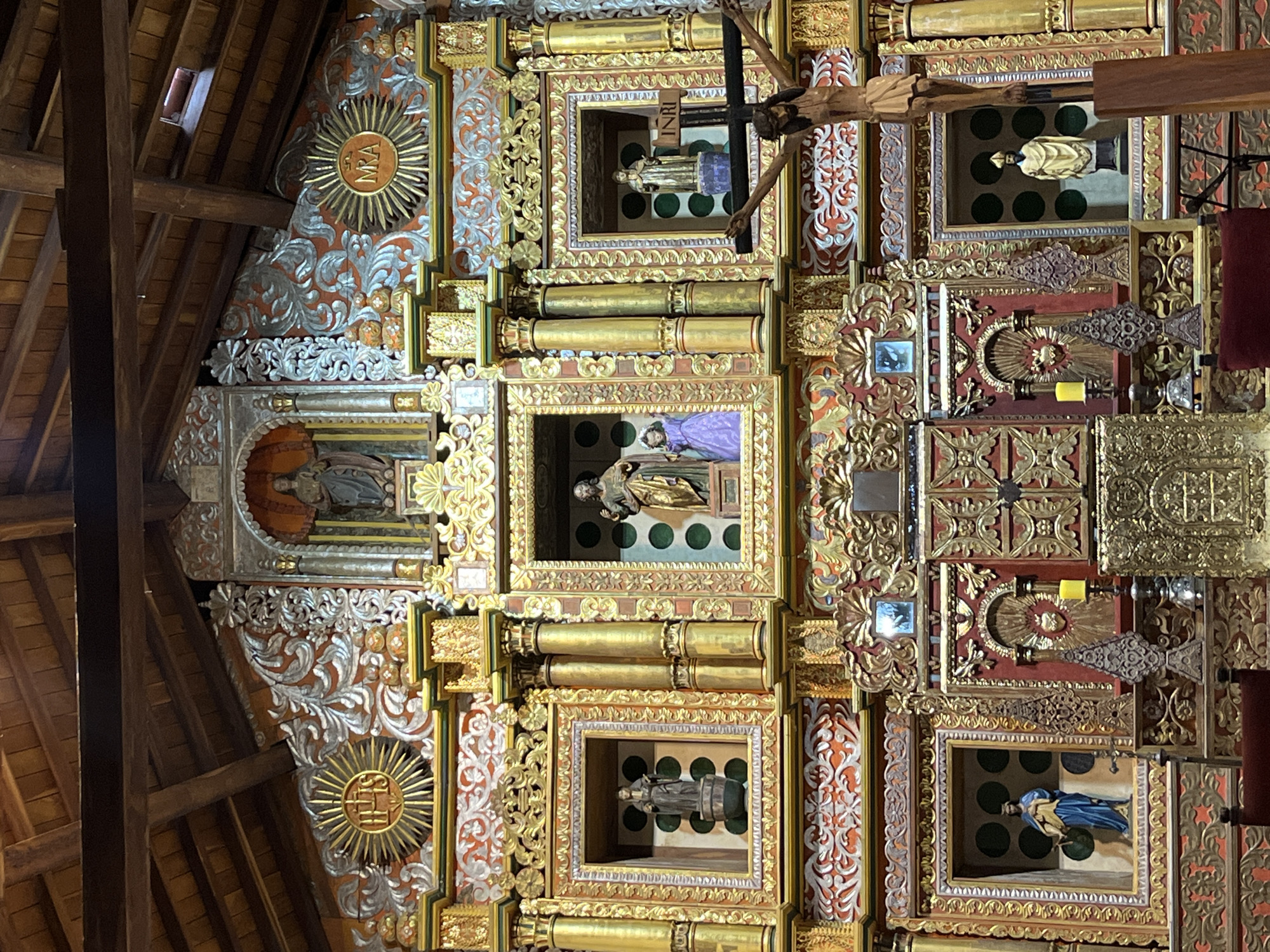 This is an interior shot of a church or religious structure. The view is angled upwards, showcasing the ornate, heavily decorated ceiling. The ceiling is divided into rectangular sections, each containing a framed religious figure or scene. The frames are gilded and embellished with intricate designs, gold columns, and floral patterns. The background of these framed sections appears to be painted with green dots. A wooden cross with a figure of Christ hangs among the framed religious figures. The ceiling is supported by wooden beams. On the left side of the image, there is decorative scrollwork painted in silver and reddish-brown tones. The scrollwork features a golden sun-like emblem with a monogram within it. The overall style and decor suggest a Baroque or Rococo influence, indicating a historic or traditional religious building.