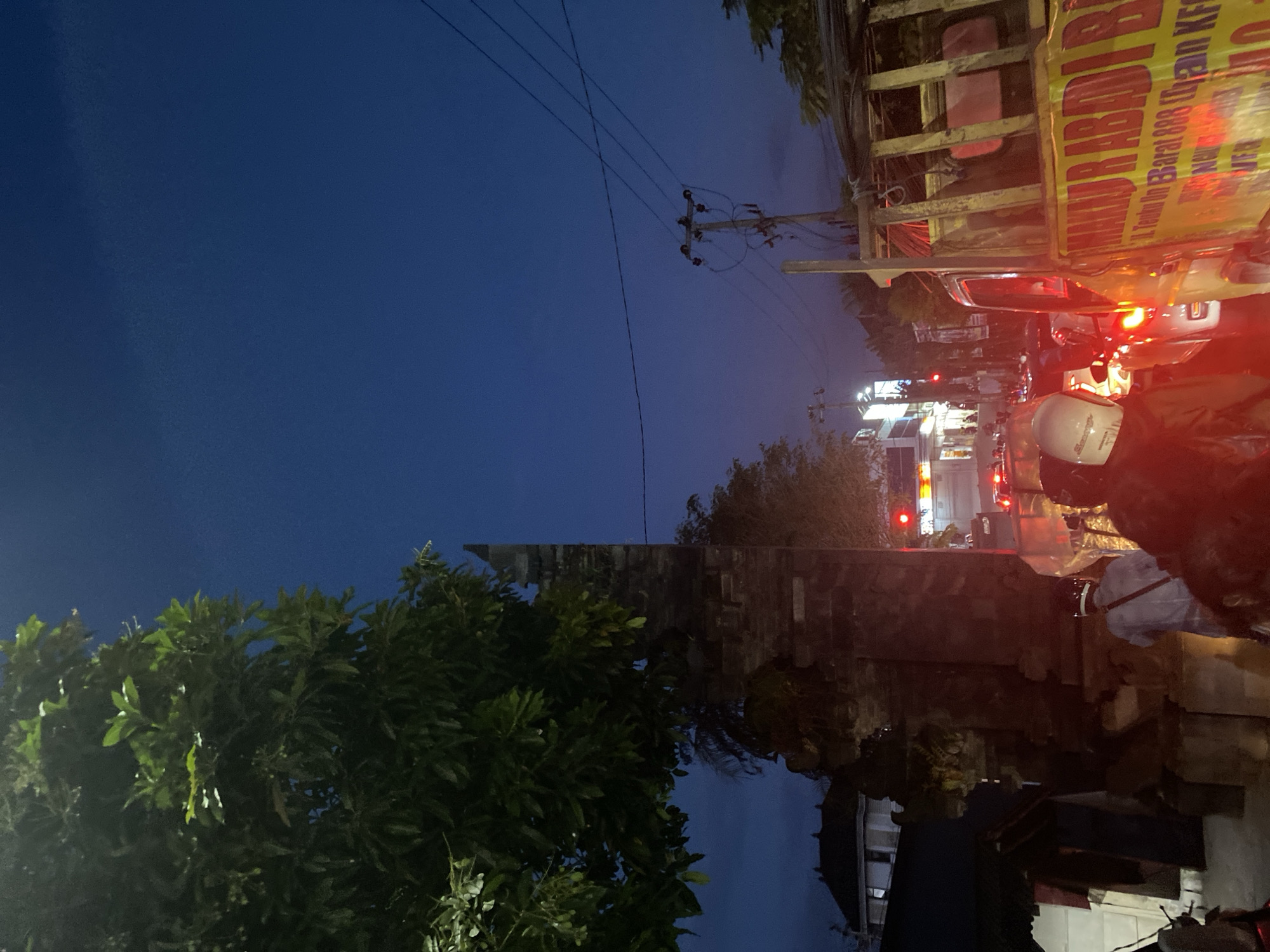 The image captures a street scene at night in North Kuta, Indonesia. The sky is a deep blue, suggesting dusk or early evening. The perspective is slightly tilted.
On the right side of the image, there is heavy traffic. Tail lights of several vehicles are visible, indicating congestion. One vehicle displays a sign that reads "HAMAD ABDI BI," partially obscured, with other text in Indonesian. A person wearing a motorcycle helmet is prominent in the foreground, suggesting they are part of the traffic.
On the left, a lush green tree is visible. Behind the tree is a brick structure, possibly a wall or building facade. Electrical lines stretch across the upper part of the frame.
The scene suggests a typical evening commute or busy street in a residential or commercial area of North Kuta.