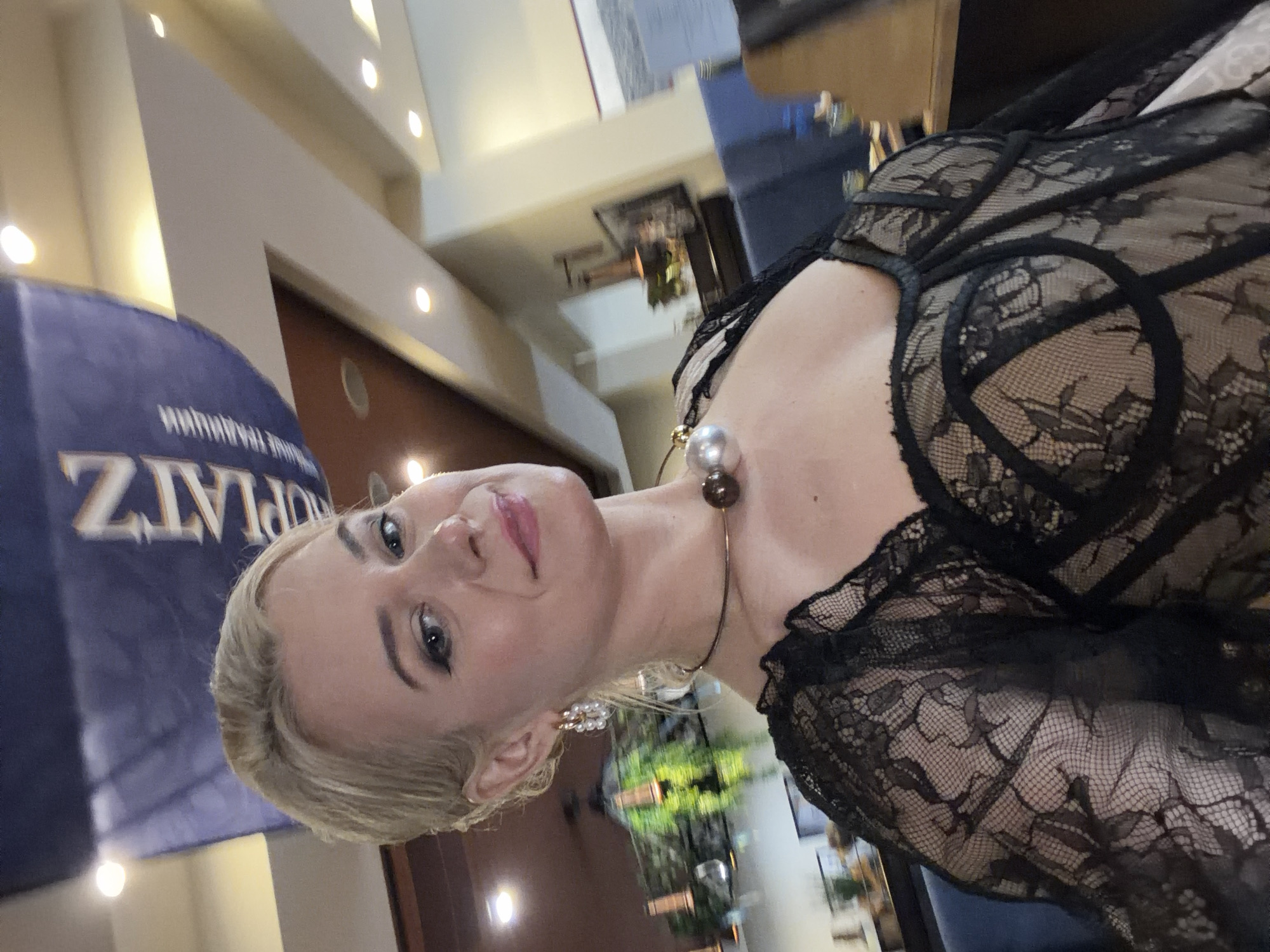 The image shows a close-up, vertically oriented, shoulder-up shot of a blonde woman with blue eyes and pink lipstick. She is wearing a black lace garment with ornate detailing around the décolletage and straps. She has a large, pearl-like earring in her left ear and a choker necklace with a dark, orb-like pendant. The background is blurred, but appears to be an indoor setting with warm lighting. There's a hint of a blue banner with white text to the left of the woman, and some greenery is visible in the reflection of a surface behind her. The woman's gaze is directed towards the camera with a slight smile. The overall mood seems to be elegant and possibly festive.