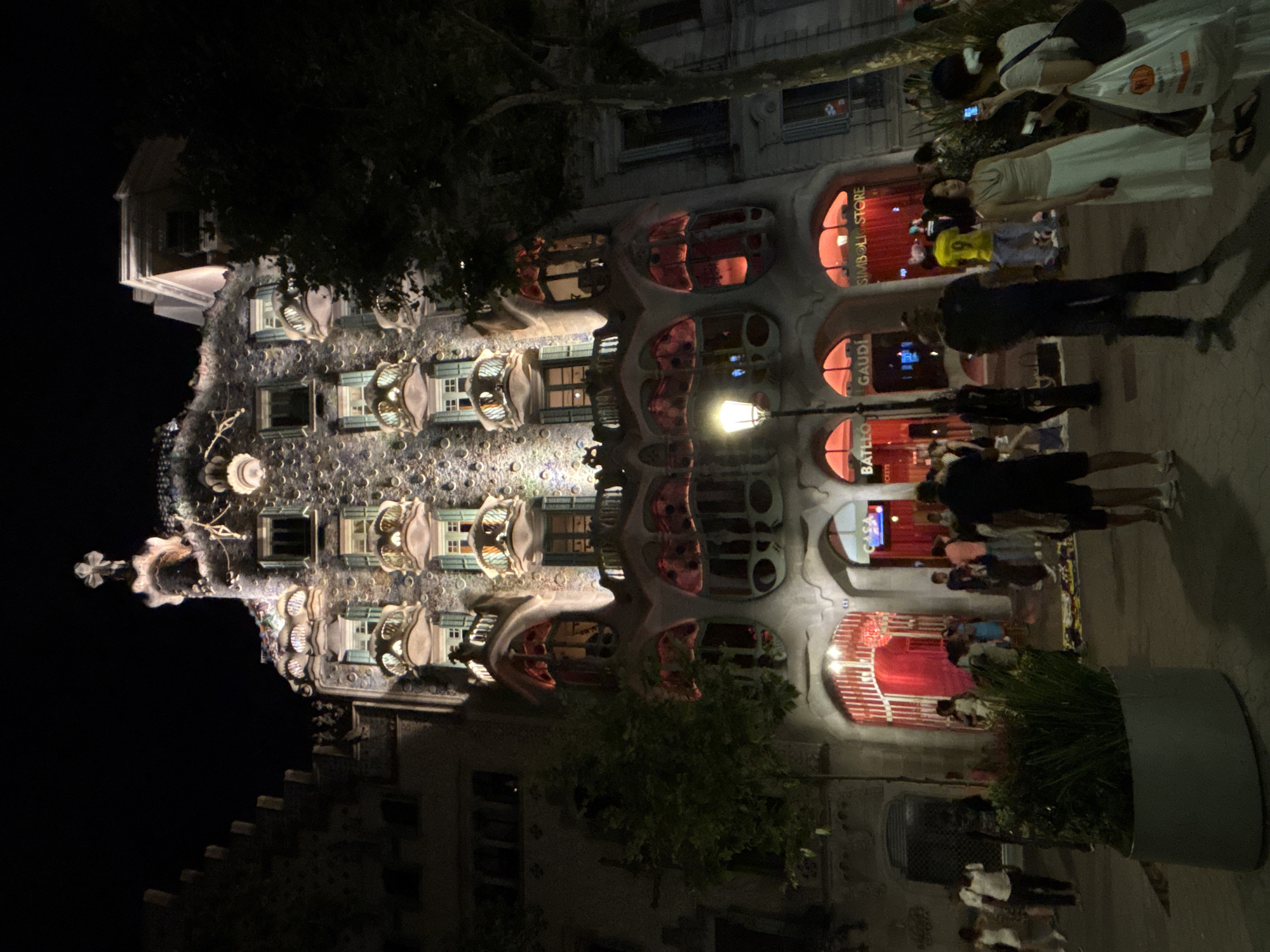 This is a nighttime outdoor photograph of Casa Batlló, a building designed by Antoni Gaudí, located on Passeig de Gràcia in Barcelona, Spain. The building's facade is illuminated, highlighting its unique organic and whimsical architectural features, including mosaic tiles, bone-like balconies, and a dragon-like roof. Several trees are visible on the right side of the building. In front of the building, there is a sidewalk with numerous people walking, standing, and congregating. Some individuals are looking at the building, while others are engaged in conversation or taking photographs. A streetlamp is visible on the left side of the sidewalk. The ground is paved, and there is a large planter with greenery in the foreground. Shop signs, including "BATLLÓ," "GAUDÍ," and "SIMBOLI STORE," are visible at the ground level of the building. The overall atmosphere suggests a popular tourist destination, with many visitors present in the evening.