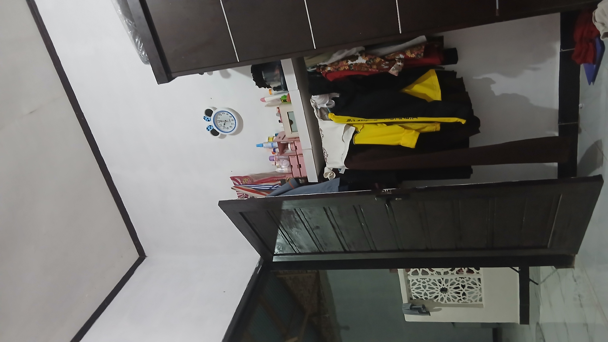 This is a medium-angle, interior shot of what appears to be a cluttered bedroom or closet area. The scene is lit by artificial light, creating shadows and highlighting textures. On the right side of the frame, a dark brown wardrobe is visible, its doors closed. To the left of the wardrobe, a shelf is mounted on the wall, displaying various items including small bottles, what look like makeup or cosmetic containers, and a blender. Above the shelf, a round clock with a Mickey Mouse design is hung on the wall. A dark brown wooden door, partially open, occupies the left side of the image. Through the doorway, another room is partially visible, showing a tiled floor, a white lattice-like partition, and what seems to be a sloped ceiling with exposed beams, possibly indicating a common Indonesian architectural style. Clothes are hung on a rack in front of the wardrobe, with a bright yellow shirt being particularly noticeable. The floor is tiled with a light marble pattern. The wall is painted white, and the ceiling is also white with dark trim. The overall impression is of a lived-in space with items stored and displayed casually.