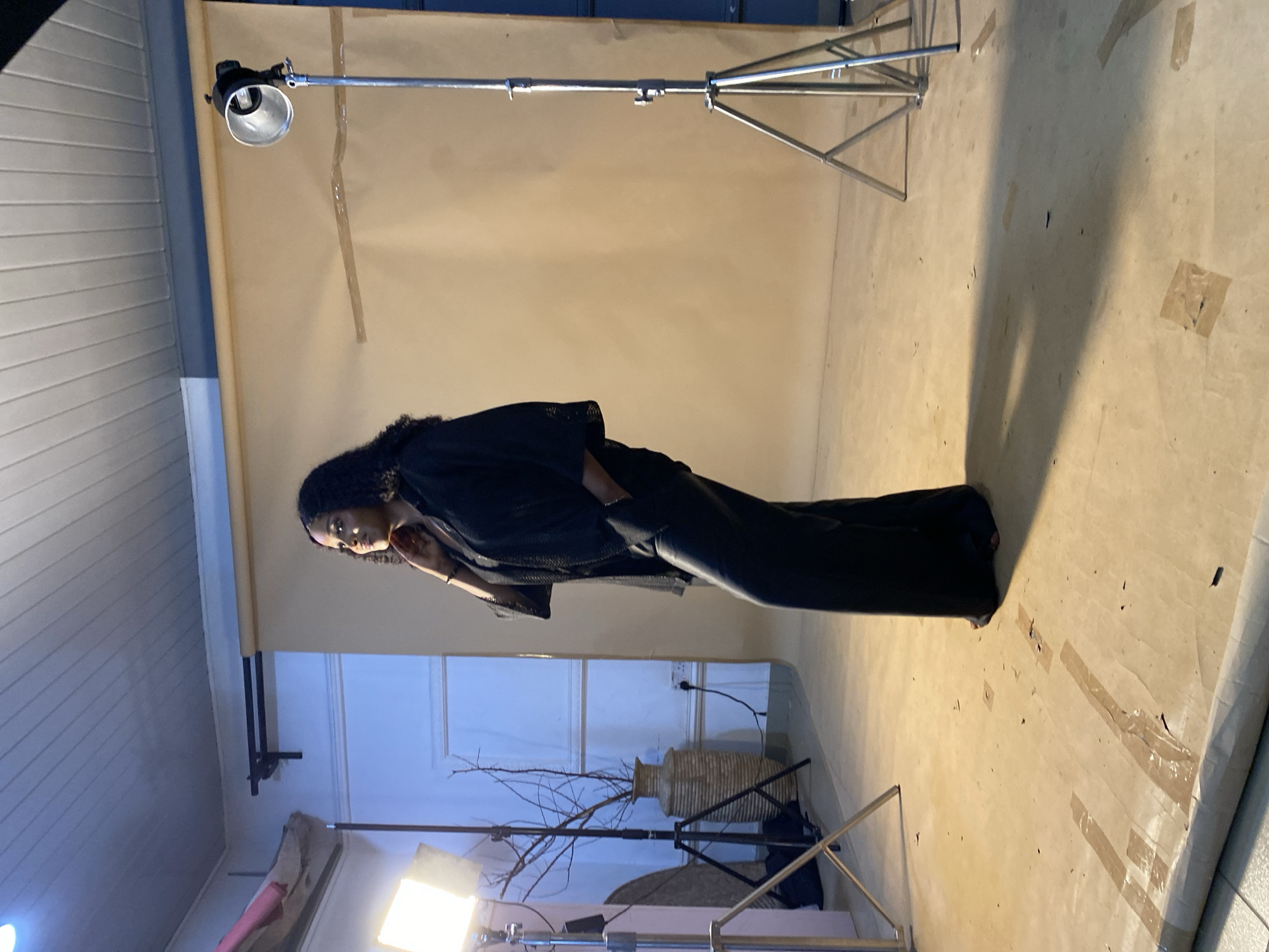 A woman is posing for a photo shoot in a studio. She is wearing a black outfit and has her hand on her chin. There are two lights set up on stands, and a backdrop of brown paper behind her. The floor is a light brown color and appears to be concrete. There is a vase with branches in the background, and a tripod with a light on it to the right of the woman. The scene is illuminated by artificial lighting.