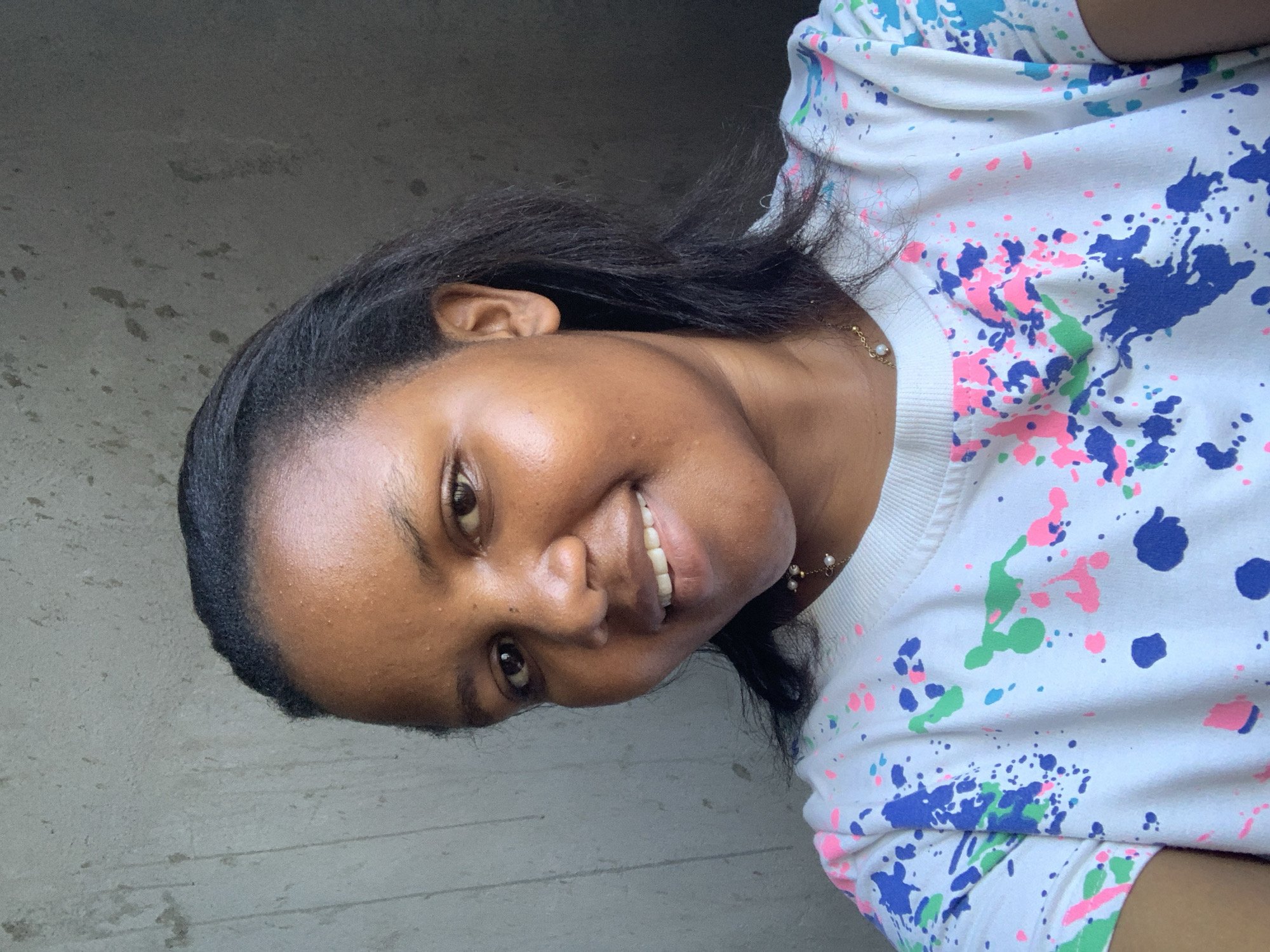 A woman is seen smiling. She has brown skin and dark hair. She is wearing a white shirt with blue, pink, and green paint splatters. She also wears a pearl necklace. The location is unknown.