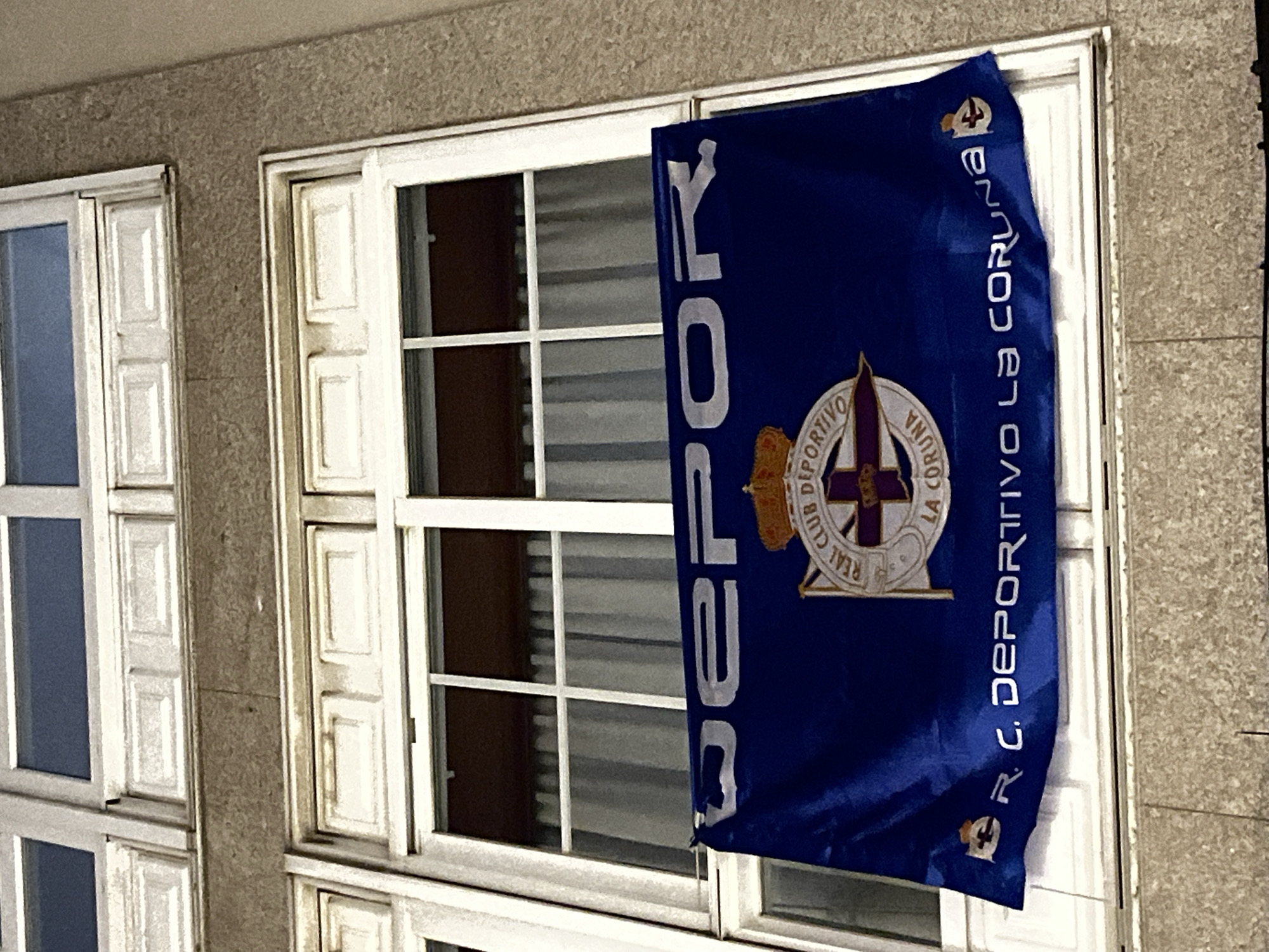The image shows the exterior of a building in A Coruña, Spain. A flag, likely a sports-related banner for R.C. Deportivo La Coruña, is hanging from one of the building's windows. The windows are white and multi-paned. The facade of the building is beige. The flag is blue with the club's emblem prominently displayed. The text "DEPOR" is visible vertically on the flag.