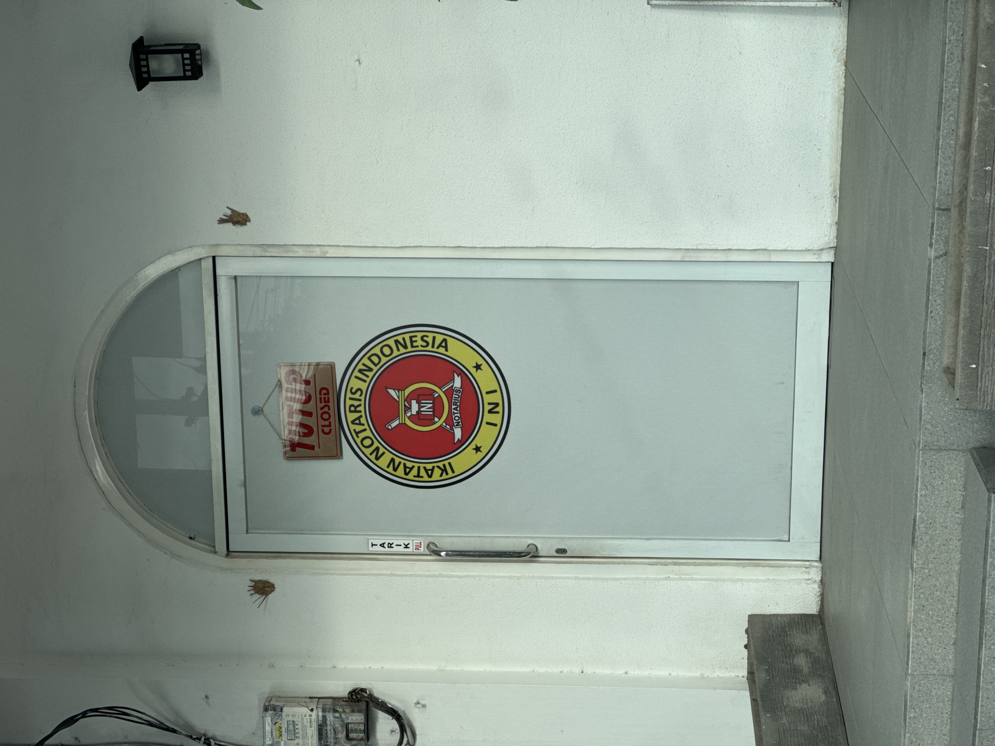 The image shows the entrance to an office in Kuta Selatan, Indonesia. The office is identified as a notary (Notaris) and is affiliated with the Indonesian Notary Association (Ikatan Notaris Indonesia). The office door is white and has a circular glass section at the top. A "CLOSED" sign is displayed on the glass. A handle with "PULL" printed in Indonesian (TARIK) is visible on the door. Above the door, there is a light fixture and a patch of what appears to be mud or dirt. Below the door, there is an electrical meter and associated wiring. A set of concrete steps is visible to the right of the door.