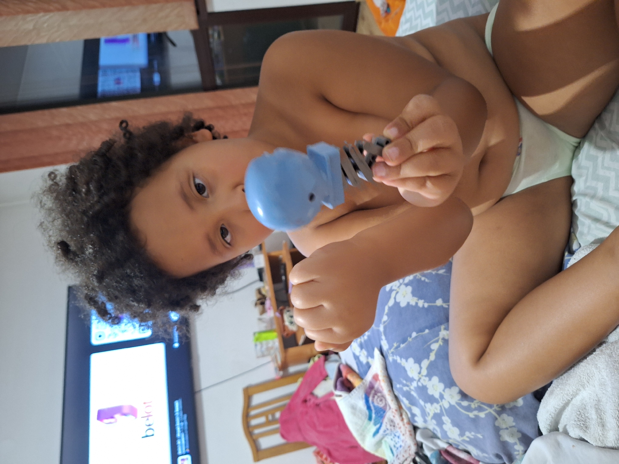 The image shows a close-up, low-angle, vertical shot of a child lying down. The child, with dark, curly hair, is wearing light-colored underwear and is holding a blue toy that resembles a whale. The child's face is visible, with eyes looking towards the camera, and an expression that could be interpreted as curious or playful. The child's legs are also prominent in the frame.
In the background, a television screen is visible, displaying content that includes the word "belot" written in pink letters. To the left of the TV, a window with blinds is visible. The setting appears to be indoors, possibly a bedroom, with a bed covered with a blue patterned blanket and a white towel. A wooden shelf unit with various items is also partially visible in the background. The lighting suggests it could be daytime, but the overall atmosphere is intimate and focused on the child and their toy. The scene is likely taking place in Blagoevgrad, Bulgaria, as indicated by the location context.