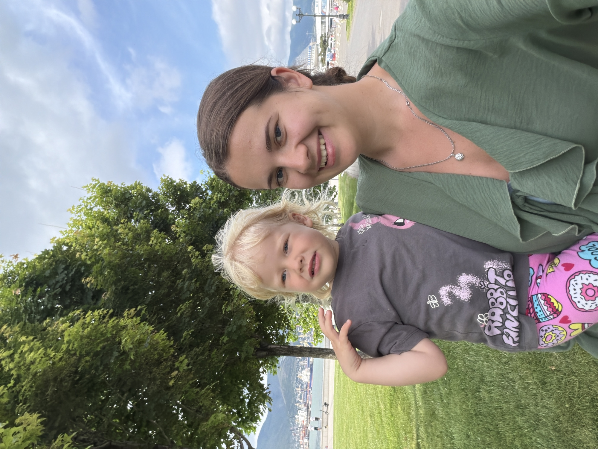 A woman and a young child are posing for a selfie outdoors, with a park and a harbor in the background. The woman, on the right, has brown hair pulled back and is wearing a green top and a silver necklace. The child, on the left, has blonde curly hair and is wearing a grey t-shirt with a graphic print and pink leggings with a donut pattern. They are standing on a grassy area, with trees to their left and a paved walkway behind them. In the distance, a harbor with ships and mountains can be seen. The sky is partly cloudy. This location is likely the embankment of the city of Novorossiysk, Russia, a coastal city on the Black Sea known for its port and scenic views.