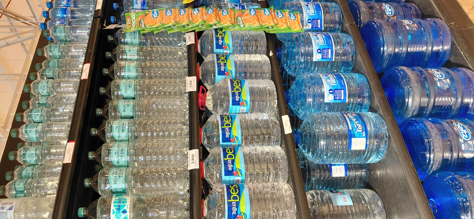 The image shows shelves stocked with bottled water in a retail environment. The top shelf displays rows of clear plastic bottles of water, many with green caps and blue or teal labels featuring text like "BENEDICTINO" and "SIN GAS." Some of these bottles appear to be 1.5-liter or similar sizes. Further to the right on the top shelf are more bottles of water, some with blue caps and labels reading "Fuente" and "Naranja." Alongside these, there is a display of individually packaged orange-flavored drinks or drink mixes, stacked vertically with labels that say "Naranja" and "10 Uds."
The middle shelf primarily features larger bottles of water with blue or green caps and labels that include "agua bes" in prominent yellow lettering. These bottles are clear plastic and appear to be of a smaller size, possibly 500ml or 1-liter. Price tags with the value "3.900" are visible on this shelf.
The bottom shelf is filled with very large, multi-gallon (likely 20-liter) water jugs. These jugs are made of clear blue plastic and have various labels. Prominent among these are jugs labeled "Torrent" and "La Fuente," both in blue and white lettering. Further to the right, more blue jugs with labels that include "Seltz" are visible.
The environment suggests a supermarket or convenience store aisle. The shelving is dark, and the background lighting is bright, with visible fluorescent light fixtures and structural elements of the ceiling. The overall impression is a well-stocked display of beverages. No people are visible in the image. The scene is consistent with a retail establishment in Encarnación, Paraguay, where such products are commonly sold.
