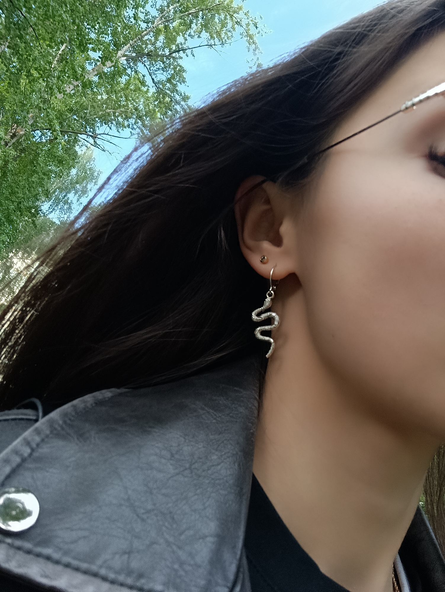 The image is a close-up, angled shot focusing on the right side of a person's head and upper torso. The individual, who has long, dark hair, is mostly seen in profile, with their right ear, neck, and shoulder prominently featured.
In the person's right ear, there are two earrings: a small, golden-colored stud in the upper lobe and a larger, silver-colored dangling earring shaped like a coiled snake in the lower lobe. The slender arm of what appears to be eyeglasses or sunglasses is partially visible near the temple area. The person is wearing a black leather-like jacket, with a round, silver-toned snap visible on the shoulder. Beneath the jacket, a collar or neckline of a black garment is visible.
The background is a vibrant outdoor scene, dominated by green tree foliage against a clear, bright blue sky. This suggests a sunny daytime setting in a park or natural area. The image captures a moment, likely a pose to showcase the unique snake earring. There is no visible text within the frame.