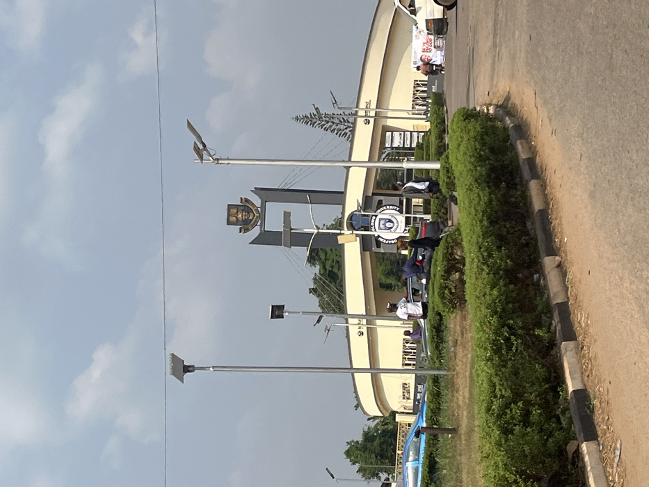 The image depicts the main entrance to Obafemi Awolowo University, located in Odan Asun, Nigeria, under a bright, partly cloudy sky. The central feature is a large, beige, curved gatehouse with a prominent dark gray archway in the center. Atop this archway is a large, gold-colored university crest, detailed with a face and an intricate shield design. Directly beneath the crest, the name "OBAFEMI AWOLOWO UNIVERSITY" is visible.
In the foreground, an asphalt road curves to the right, bordered by a central median featuring green, manicured hedges and some patches of dry grass. Multiple tall, silver-colored streetlights, each topped with a flat, rectangular solar panel, are spaced across the scene. Overhead power lines are faintly visible against the sky.
Several individuals are present near the entrance. Approximately seven people, primarily adult males, are observed. On the left side of the entrance, near a blue sedan, one person in a white t-shirt and cap stands, facing right. Another individual in a dark shirt appears to be leaning over the hood of a dark-colored sedan located in front of the entrance. Further to the right, a person in a white shirt and dark trousers stands near the roadside. On the far right, a white van is partially visible, and a person on a motorcycle or scooter displays a large white banner with red text, partially reading "BE YE TO GOD". The entrance area itself shows what appears to be a security booth or office on the left, marked with a blue circular sign. Beyond the main gate, various trees and foliage are visible.