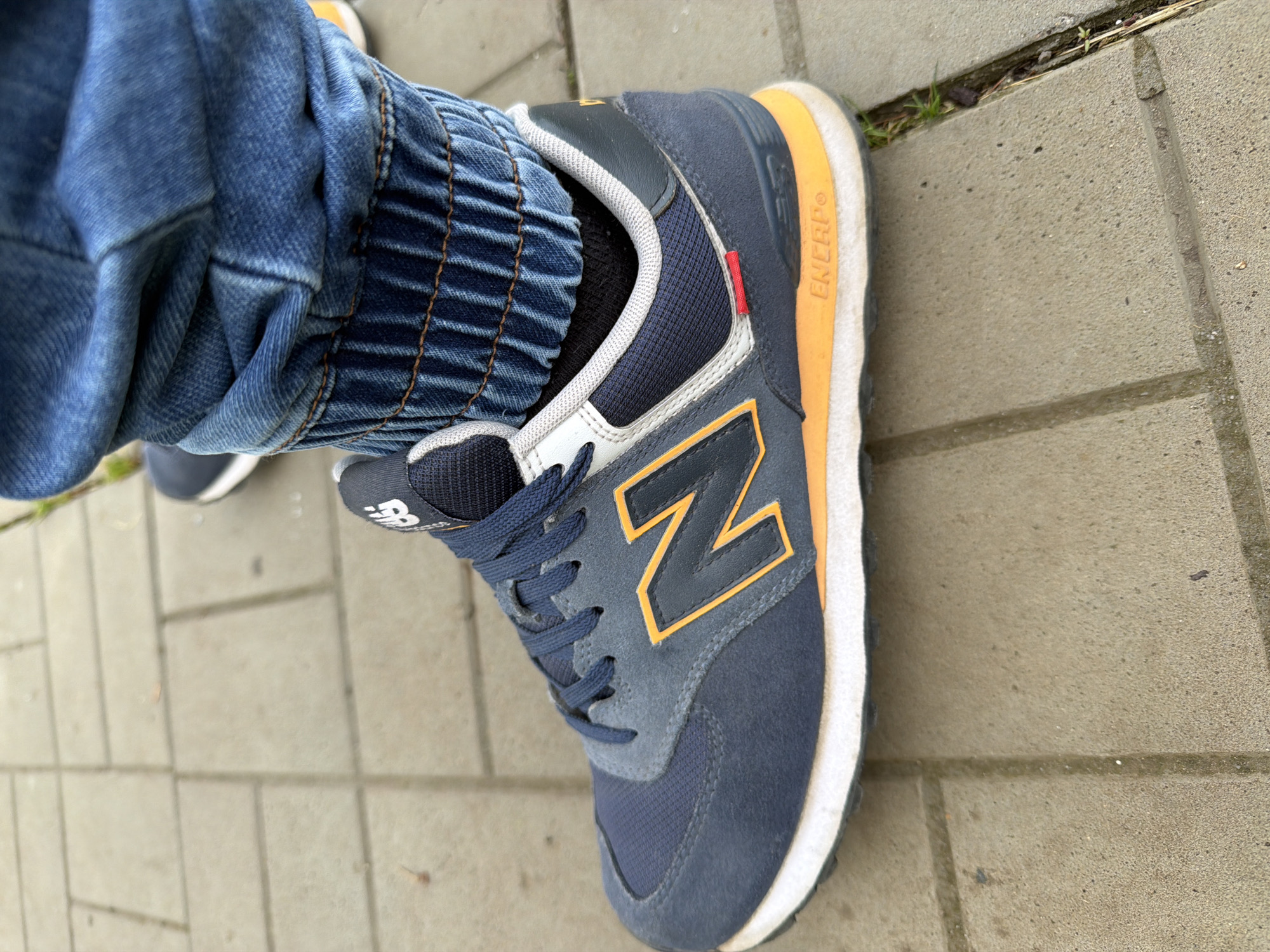 The image shows a person's left foot wearing a New Balance sneaker. The sneaker is primarily blue with a white and orange accent on the sides. The "N" logo is visible on the side of the shoe. The person is wearing denim pants with an elastic cuff around the ankle. The person is standing on a paved surface.