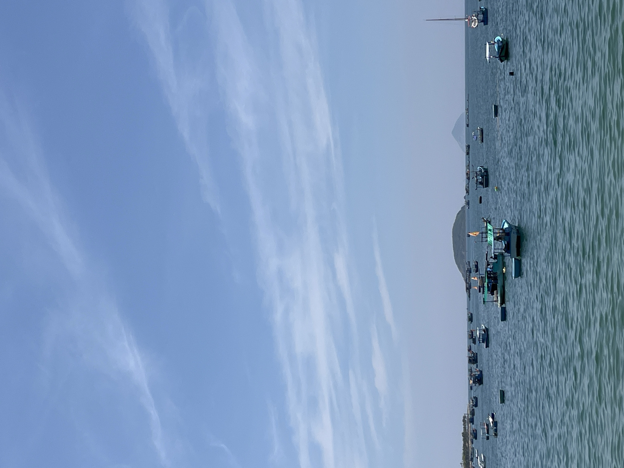 The image captures a broad coastal vista in Bac Nha Trang Ward, Vietnam, under a clear, bright blue sky containing scattered, thin white clouds. The lower half of the frame is dominated by a calm, deep blue-green body of water. Distributed across the water are numerous small to medium-sized fishing boats, primarily painted in shades of blue and green, with some white details. Several boats feature small canopies, and a few display small yellow flags. A single sailboat with a tall mast is visible on the far right. The horizon reveals landmasses; a vegetated coastline is present on the far left, and multiple distant islands or hills, including a distinct rounded peak, are silhouetted against the lighter sky. No individuals are visibly present on the boats or in the water.