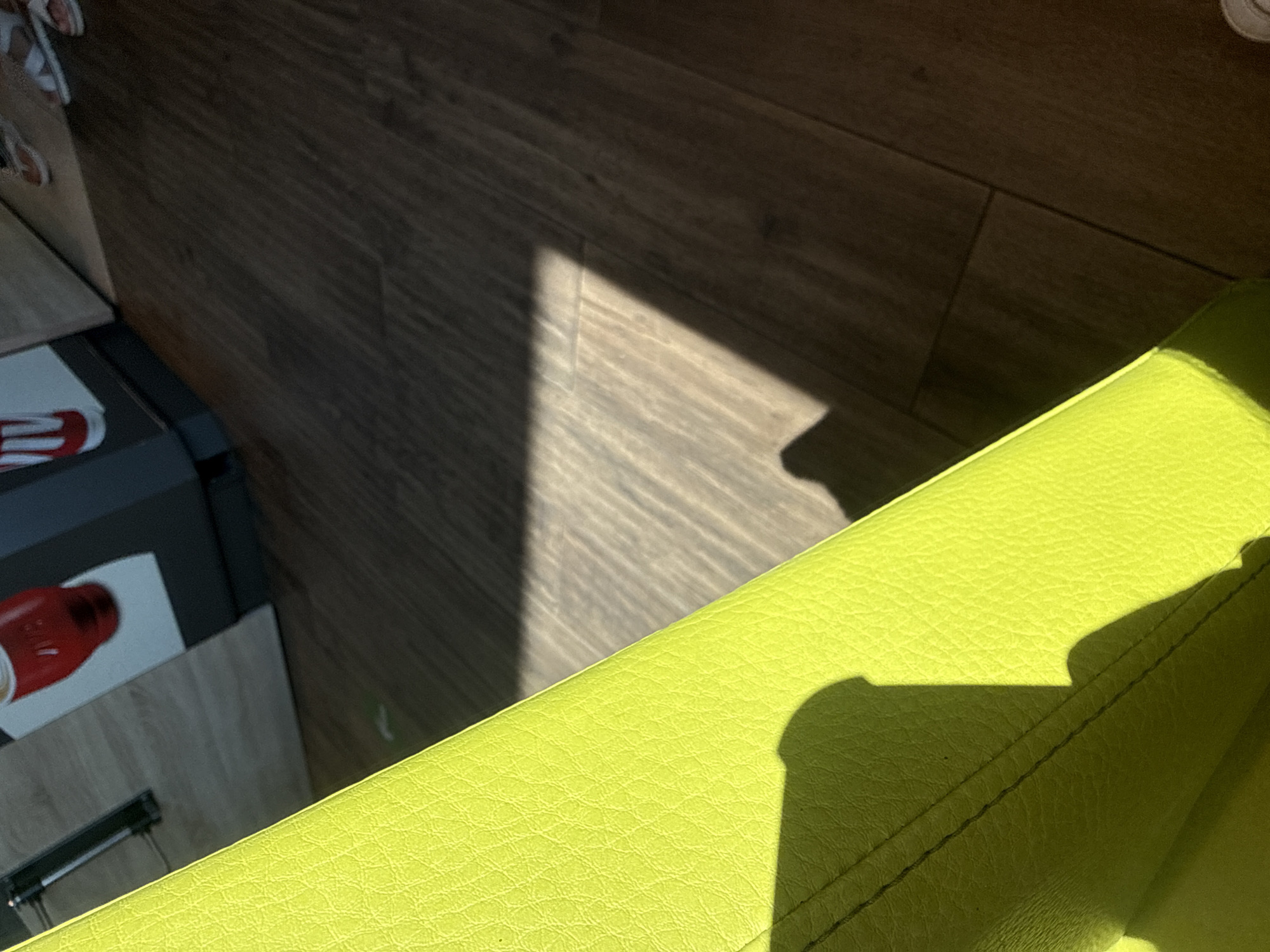 In Drazhevo, Bulgaria, an extreme close-up shot captures a bright lime-green textured surface, likely upholstery, dominating the left side of the frame. Sunlight casts a sharp, geometric shadow across a wooden floor with visible grain and plank lines on the right. In the background, out of focus, are elements of what appears to be a retail or service establishment, including a dark gray kiosk or counter with images of beverages, one red and one with red and white stripes. The floor in the background transitions from lighter wood tones to darker shades. The overall impression is an indoor setting illuminated by strong directional light.
