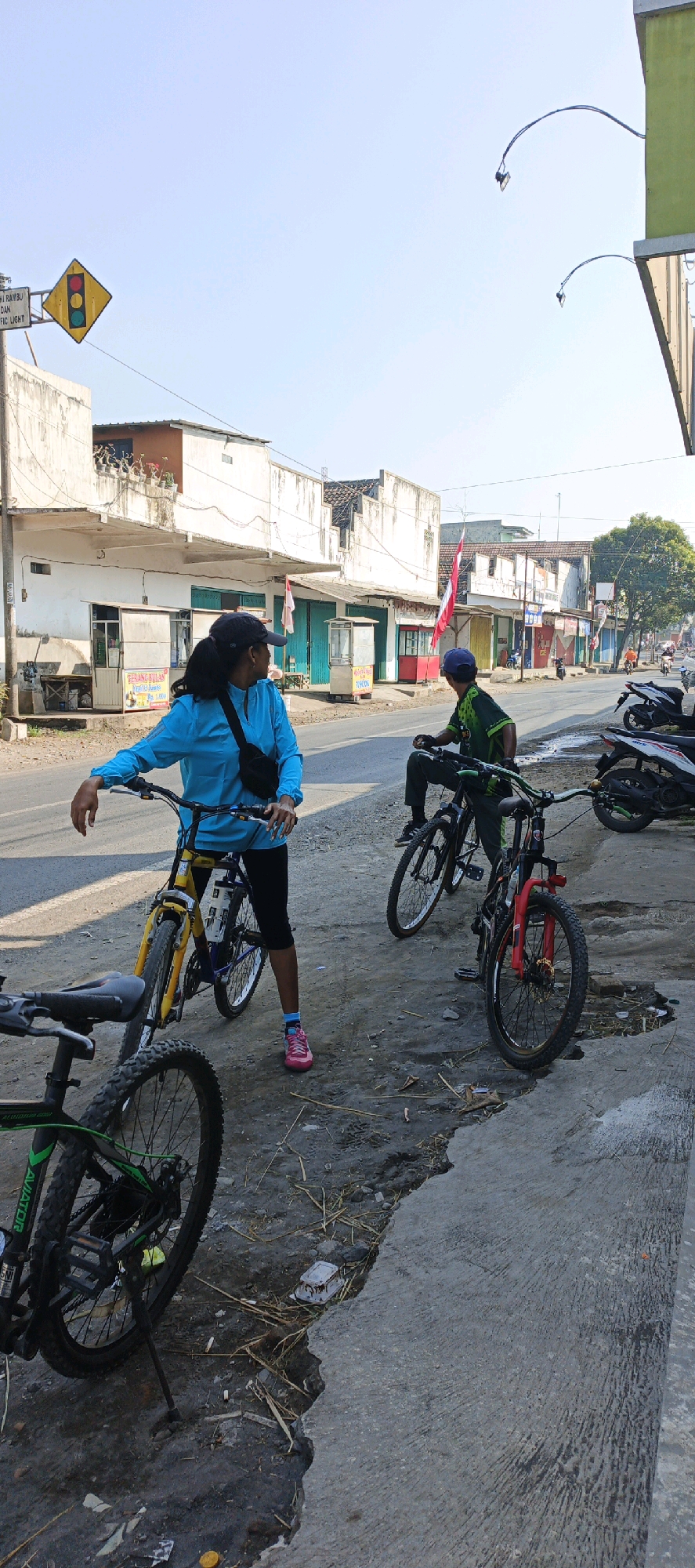 In Menampu, Indonesia, two individuals are stopped on the side of a paved road, flanked by parked motorcycles and a row of small shops. The individual on the left, wearing a bright blue long-sleeved shirt and black leggings, is standing next to a yellow and purple bicycle. Their left hand rests on the handlebars, and they are looking to the right. A black fanny pack is worn across their chest. The individual on the right, wearing a green and black cycling jersey, is seated on a black bicycle with red accents on the front fork, facing the left. They are wearing a blue cap and black cycling gloves. The environment features a clear blue sky above, with buildings lining the road. One building has a red and white Indonesian flag displayed prominently. The ground along the side of the road is uneven and appears to be a mix of dirt, gravel, and some discarded debris. A traffic light is visible on a pole in the upper left of the frame.