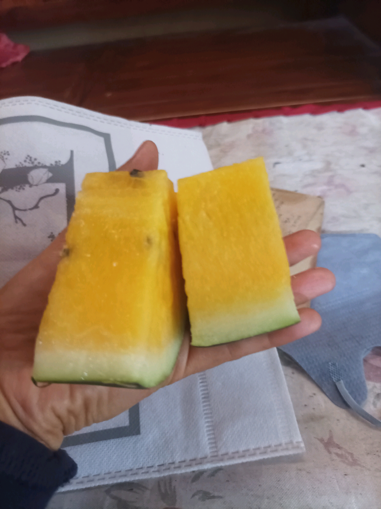 A hand is holding two slices of yellow watermelon. The watermelon slices are rectangular with a yellow flesh and a thin green rind. The hand is a light skin tone and is positioned in the center of the frame, supporting the fruit. The background shows a light-colored cloth with a pattern and a dark wooden surface. There are also some other objects in the background, including a brown paper bag and a blue object that appears to be a face mask. The lighting suggests an indoor setting, likely during the daytime. The image does not provide any specific visual cues to determine the location in Canga'an, Indonesia.