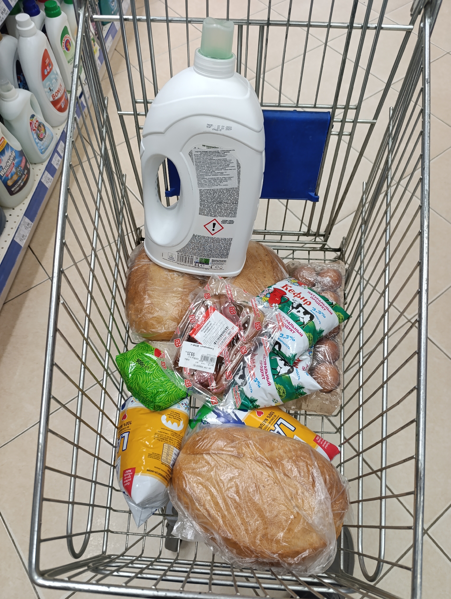 In Dubăsari town council, Moldova, a metal shopping cart is filled with groceries. Dominating the cart is a large white bottle of liquid detergent, its cap a vibrant green. Beneath it, two loaves of bread are individually wrapped in clear plastic. To the right, a package of brown eggs is visible, alongside a green package depicting a cow and the word "Кефир" (Kefir), indicating a dairy product. Further items include a green bag with white wavy lines and a yellow package with red lettering. The background consists of a tiled floor and shelves stocked with various cleaning products, partially visible on the left side. The scene is captured from a top-down perspective, focusing on the contents of the shopping cart.