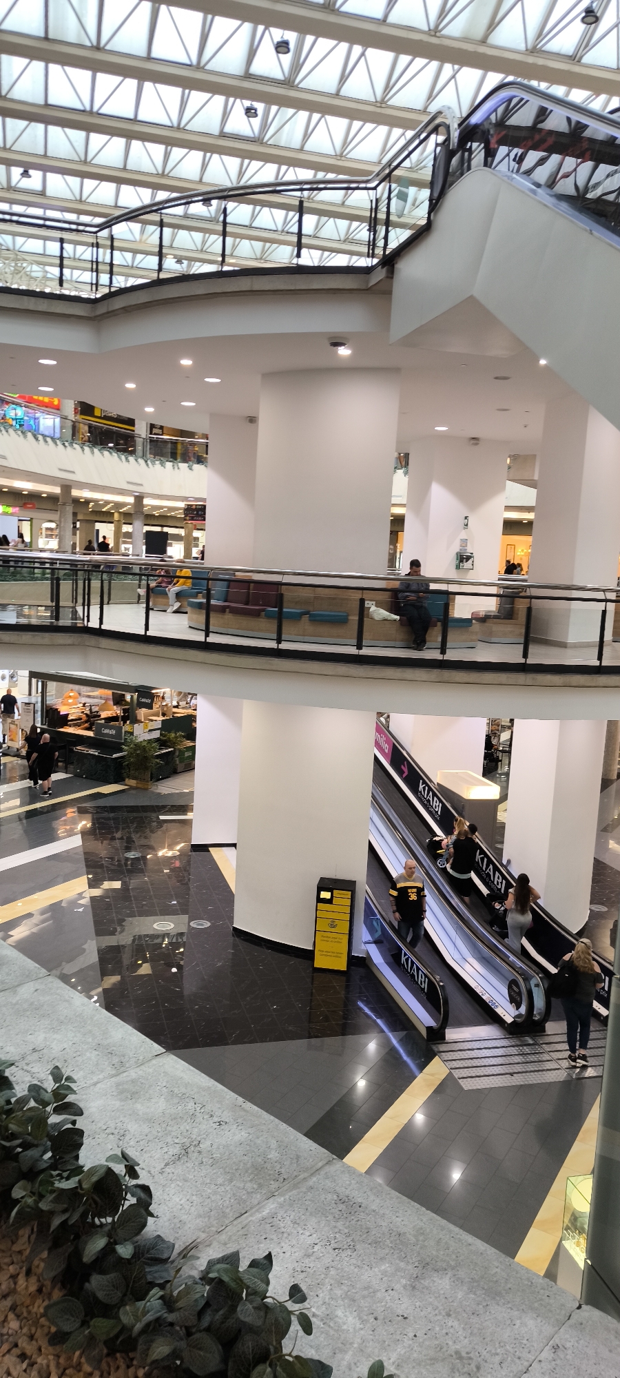 This vertical shot captures the multi-level interior of a brightly lit shopping mall, likely in Barberà Del Vallès, Spain. The upper part of the image shows a modern, geometric skylight ceiling constructed with glass panels and a white metal framework, allowing natural light to illuminate the space.
Below the skylight, the mall's architecture features curving balconies with sleek glass and black metal railings, supported by large, cylindrical white pillars. On an intermediate level, several people are visible. Some individuals are seated on integrated bench seating with blue and purple upholstered sections, with one man in a dark hoodie appearing engrossed in his phone. This area overlooks lower levels where more shops and activity can be seen.
The lower main floor is characterized by dark, highly reflective marble-like tiles with lighter beige or yellow geometric patterns, mirroring the overhead lighting. Two downward-moving escalators dominate the right side, both prominently displaying "KIABI" branding. One escalator also shows a partial word, "amilia," accompanied by an arrow. People are using the escalators, including a man in a yellow and black jersey with the number "36" and a woman with a stroller.
To the left on the lower level, a cafe area is visible, identified by a "Cafécafé" sign. Staff can be seen behind a counter. Near the escalators, a distinctive yellow and black kiosk or service station stands. In the immediate foreground, part of a concrete planter with artificial green foliage and light-colored pebbles adds a decorative element. The scene appears to be active but not overly crowded, consistent with a typical shopping mall environment during the day.