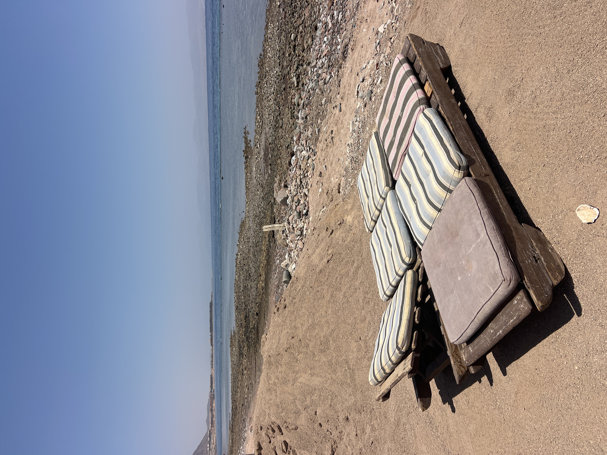 In Nueiba, Egypt, a rustic, weathered wooden lounger with several cushions sits on a sandy beach. The lounger is constructed from what appears to be reclaimed wood, possibly pallets, and shows signs of age and exposure to the elements. Multiple cushions, some with striped patterns in blues, yellows, and pinks, and one solid grey cushion, are arranged on the lounger. The background features a rocky shoreline leading to a calm, blue body of water. Distant mountains and a hazy horizon complete the serene beach scene.