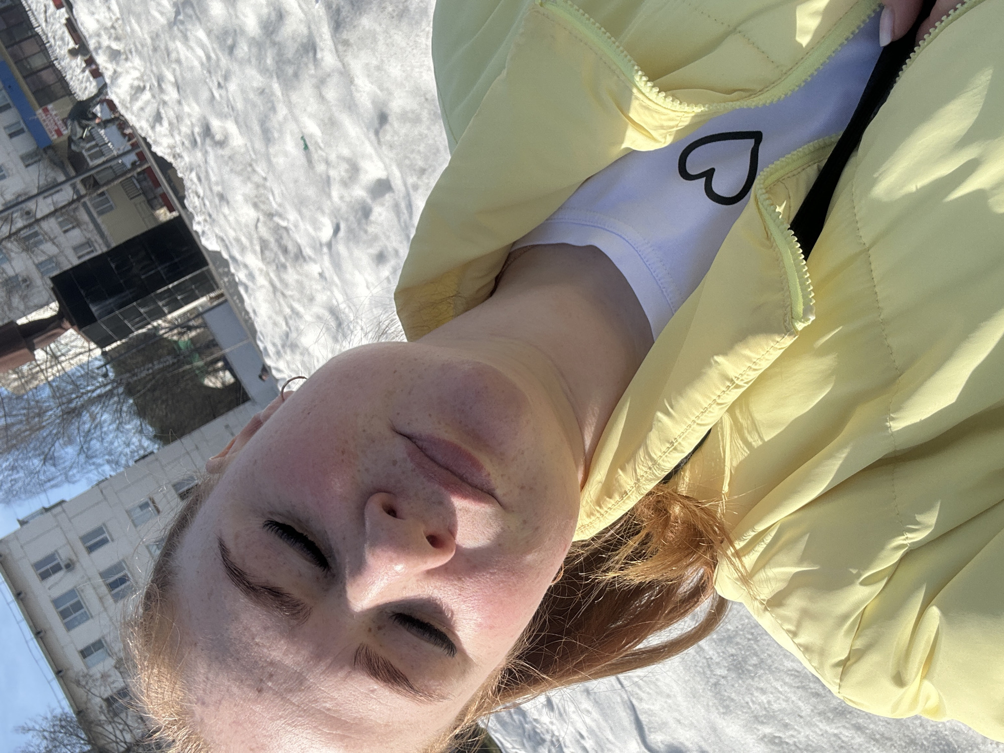 A fair-skinned young adult female lies supine on a snow-covered surface in what appears to be an urban environment. Her eyes are closed, and her freckled face is turned slightly upward. She wears a white t-shirt with a black heart graphic under an open, light yellow padded jacket. In the background, blurred images of buildings suggest a residential area. The presence of snow indicates winter. The image is oriented in portrait mode. Based on the image, determining the specific location in Chaikovsky, Russia, is not possible.