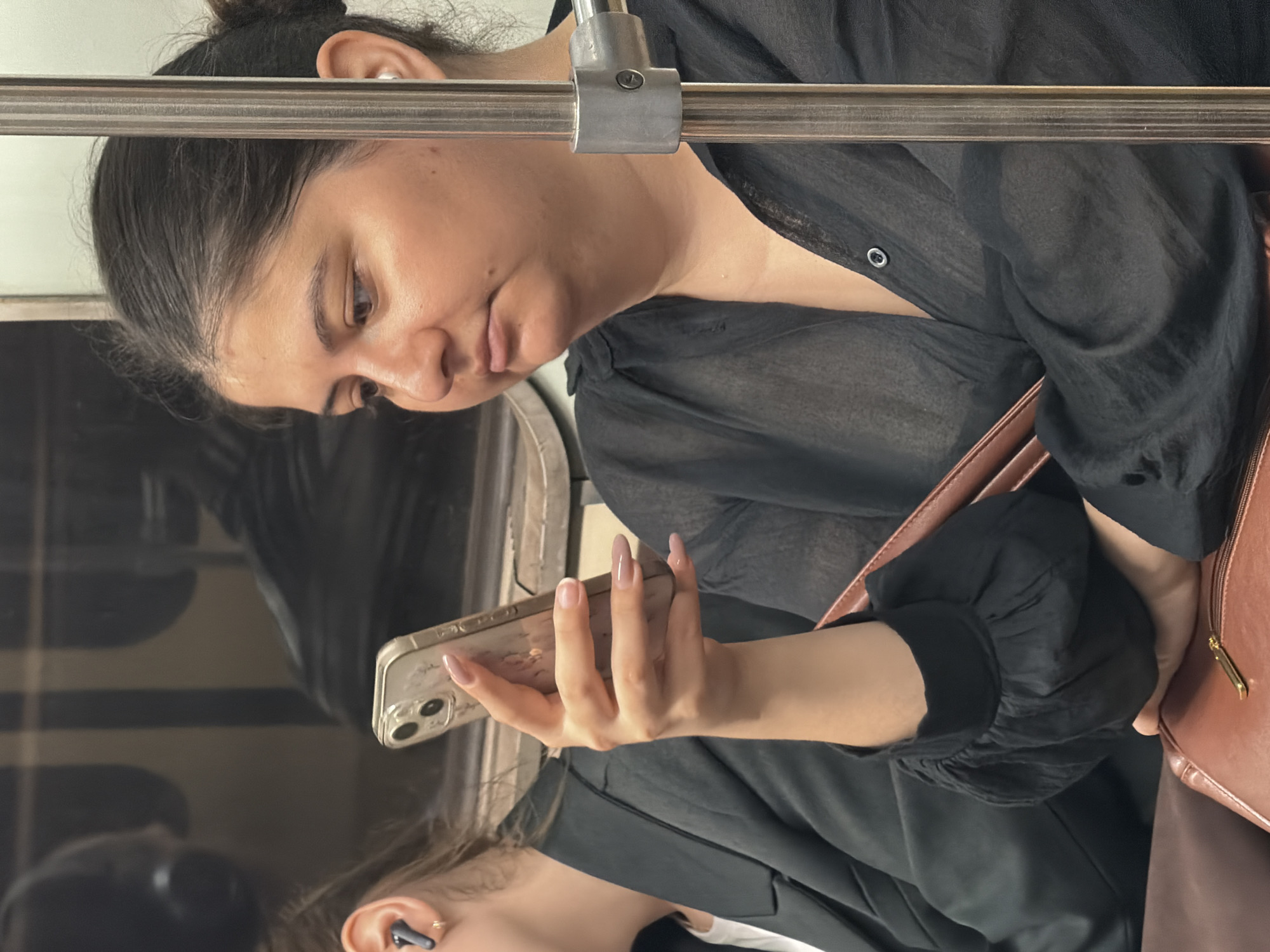The image shows two individuals inside a subway car, likely in Moscow, Russia, given the context. The primary subject, a woman with dark hair pulled back and wearing a sheer black button-up shirt, is seated and looking down at her mobile phone, which she holds in her right hand. Her fingernails are painted a light pink. Next to her, a second individual, partially visible and wearing a dark blazer over a white top, has an earbud in their left ear. The interior of the subway car is dimly lit, with visible metal poles and seating. The overall scene depicts a mundane moment of daily commute.