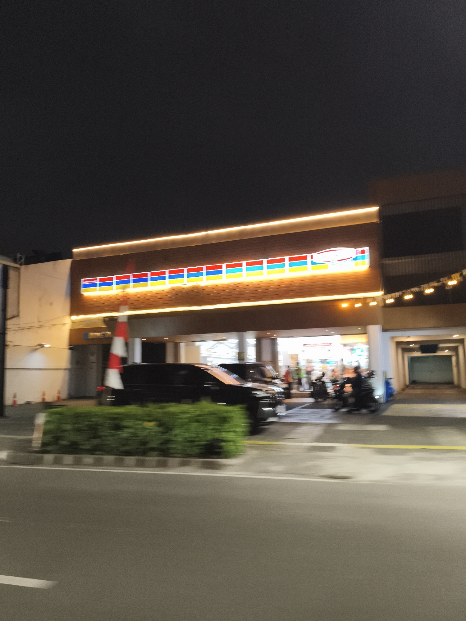 The image depicts a night scene of a brightly lit convenience store with its characteristic red, yellow, and blue striped logo prominently displayed on a wooden facade. The store appears to be a retail establishment located on a street in South Jakarta, Indonesia, as indicated by the presence of a red and white striped pole, possibly a flag or marker.
In front of the store, several vehicles are visible. A black SUV is parked to the left, and in the center, a group of people are gathered, some on motorcycles. A white and red striped pole is positioned near the entrance of the store, and orange traffic cones are lined up along the sidewalk.
The environment suggests an urban setting, with a paved road in the foreground and buildings in the background. The lighting is artificial, with the store's sign and exterior lights illuminating the area. The overall impression is that of a bustling commercial street at night.