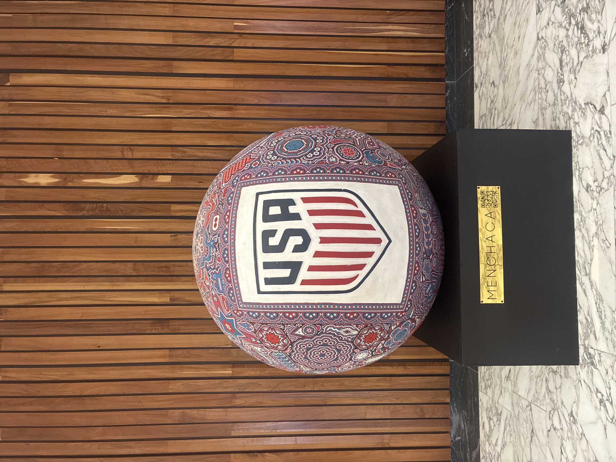 The image shows a patterned sphere, presumably a decorative object, prominently displaying the USA logo. It rests on a wood-planked floor. Next to the sphere is a black rectangular box with a gold plaque that reads "MENCHACA" and includes a QR code. The box is partially situated on a floor of white and gray marble. The overall setting suggests an interior space with a modern design. Based on the text on the plaque, it is possible this is located in Mexico City.