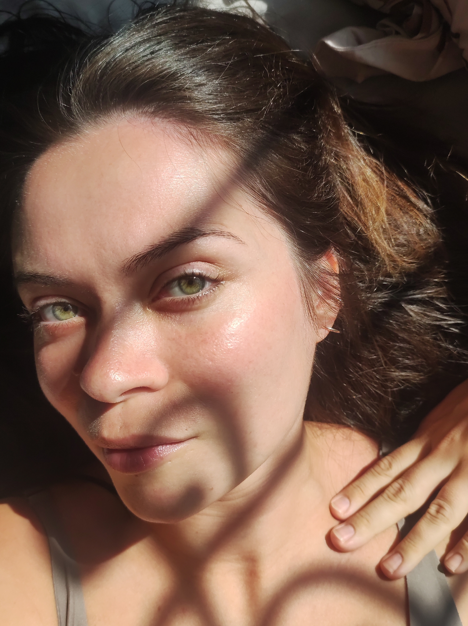 The image is a close-up, high-angle selfie of a woman lying down. She has long, dark brown hair that is slightly wavy and falls around her face and shoulders. Her eyes are green and she is looking directly at the camera. The lighting suggests she is outdoors, possibly near a window, as a patterned shadow is cast across her face and chest. She is wearing a gray tank top. Her right hand is resting on her collarbone and neck. The environment appears to be indoors, based on the presence of what looks like bedding or fabric in the background. There is no information in the image that would indicate a specific location within the city of Chaikovsky, Russia.