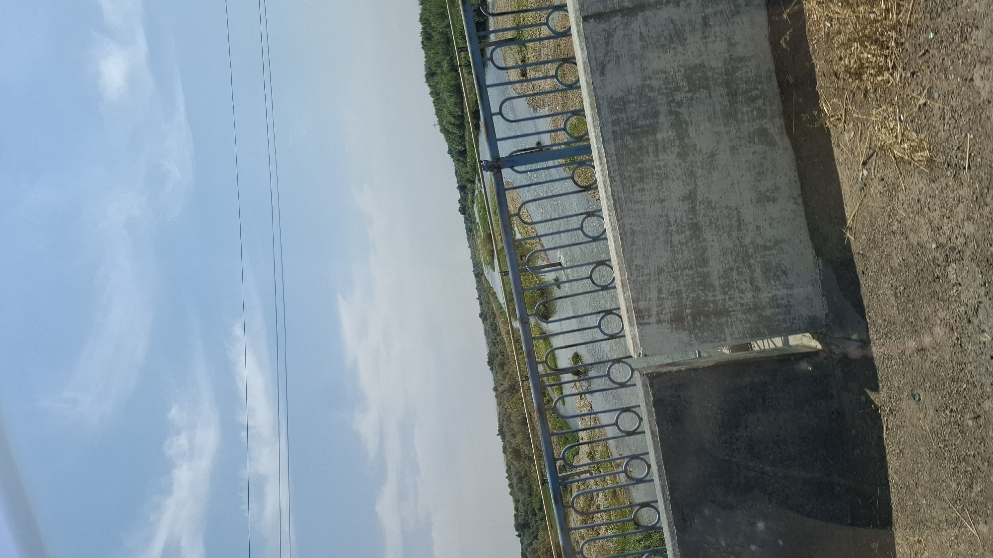 This is a vertical, eye-level, medium shot of a river as seen from a bridge. The sky is a light blue with scattered clouds. In the foreground of the image is a concrete barrier and a metal railing. Beyond the railing, a river flows with visible banks covered in pebbles and vegetation. The river is relatively narrow and appears to be in a rural or semi-rural area, with trees and greenery lining the banks in the distance. The scene is brightly lit by sunlight. The location is identified as a bridge in the city of Құтарыс, Kazakhstan. There are no people or animals visible in the image.