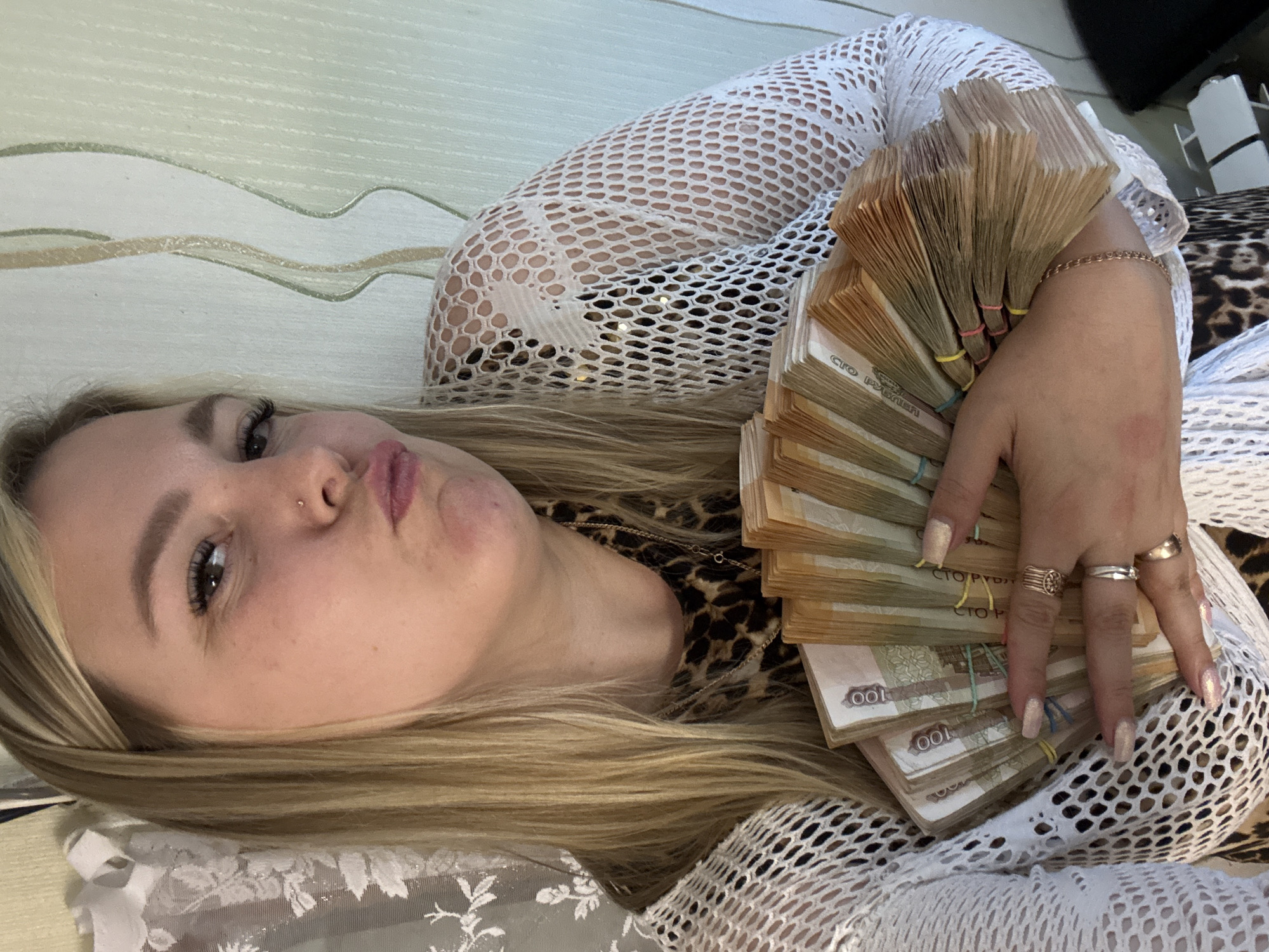 A woman is lying down, holding a large amount of Russian Ruble banknotes in her hand. She is wearing a white, net-like top and a leopard-print garment underneath. Her blonde hair is spread out on the surface beneath her. The background includes a wall with a wavy design. The location is implied to be in the city of Noviy, Russia, based on the currency.