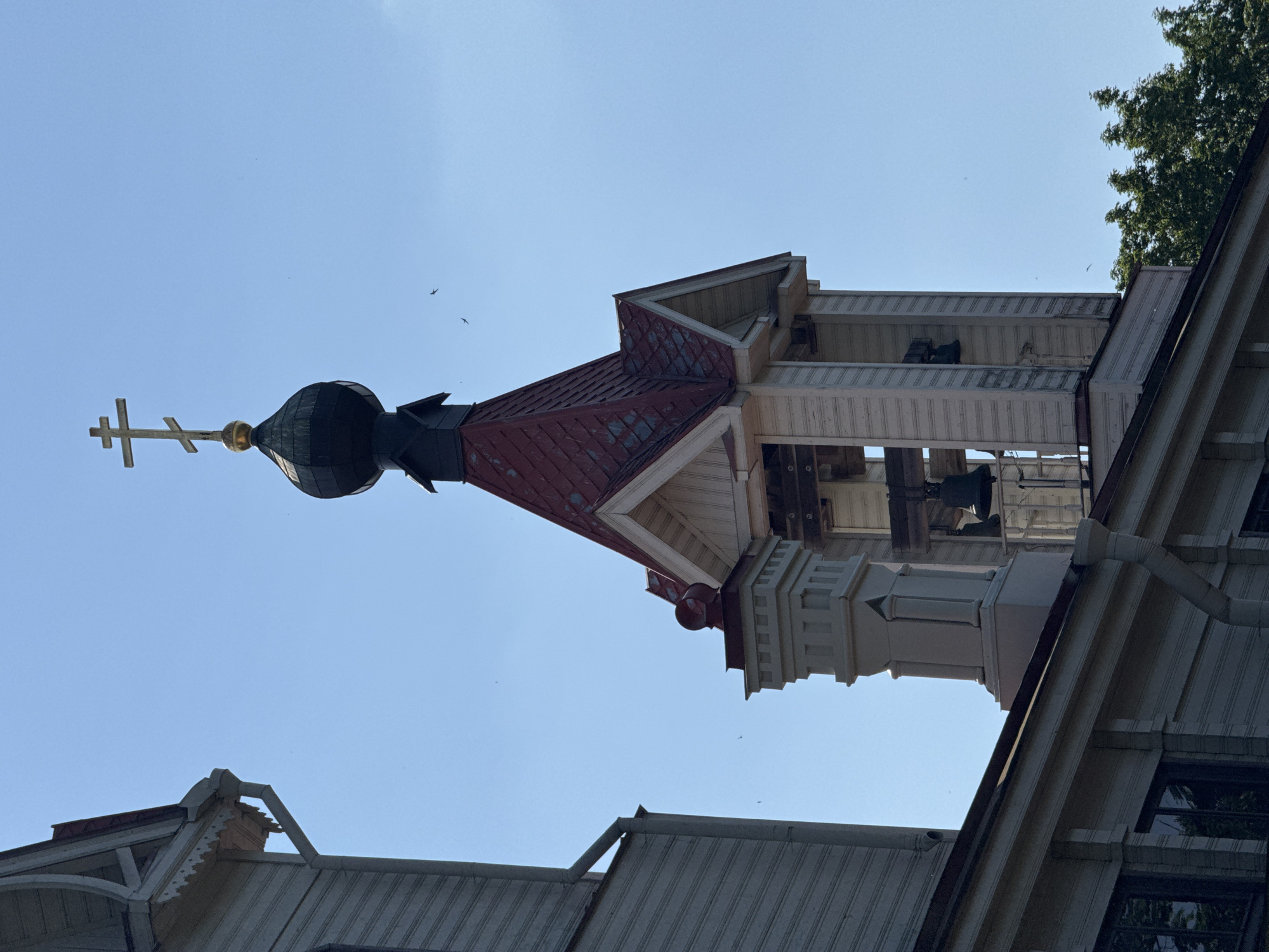 The image depicts the upper sections of an Orthodox church against a clear blue sky. The central subject is a bell tower with a red, multi-faceted roof, adorned with a golden cross at its peak. Below the cross, a dark, onion-shaped dome is visible. The bell tower features wooden latticework and bells hanging within its open structure. To the right, a portion of the church's main building is visible, characterized by light-colored siding and dark-framed windows. In the foreground and to the left, sections of other church roofs, with corrugated metal and decorative trim, are visible. Several small birds are flying in the sky. The lighting suggests it is daytime, likely morning or afternoon, with clear weather. The image is oriented vertically, making it appear as if the church is ascending into the sky. No text is visible in the image. The location is Lomonosov, Russia.