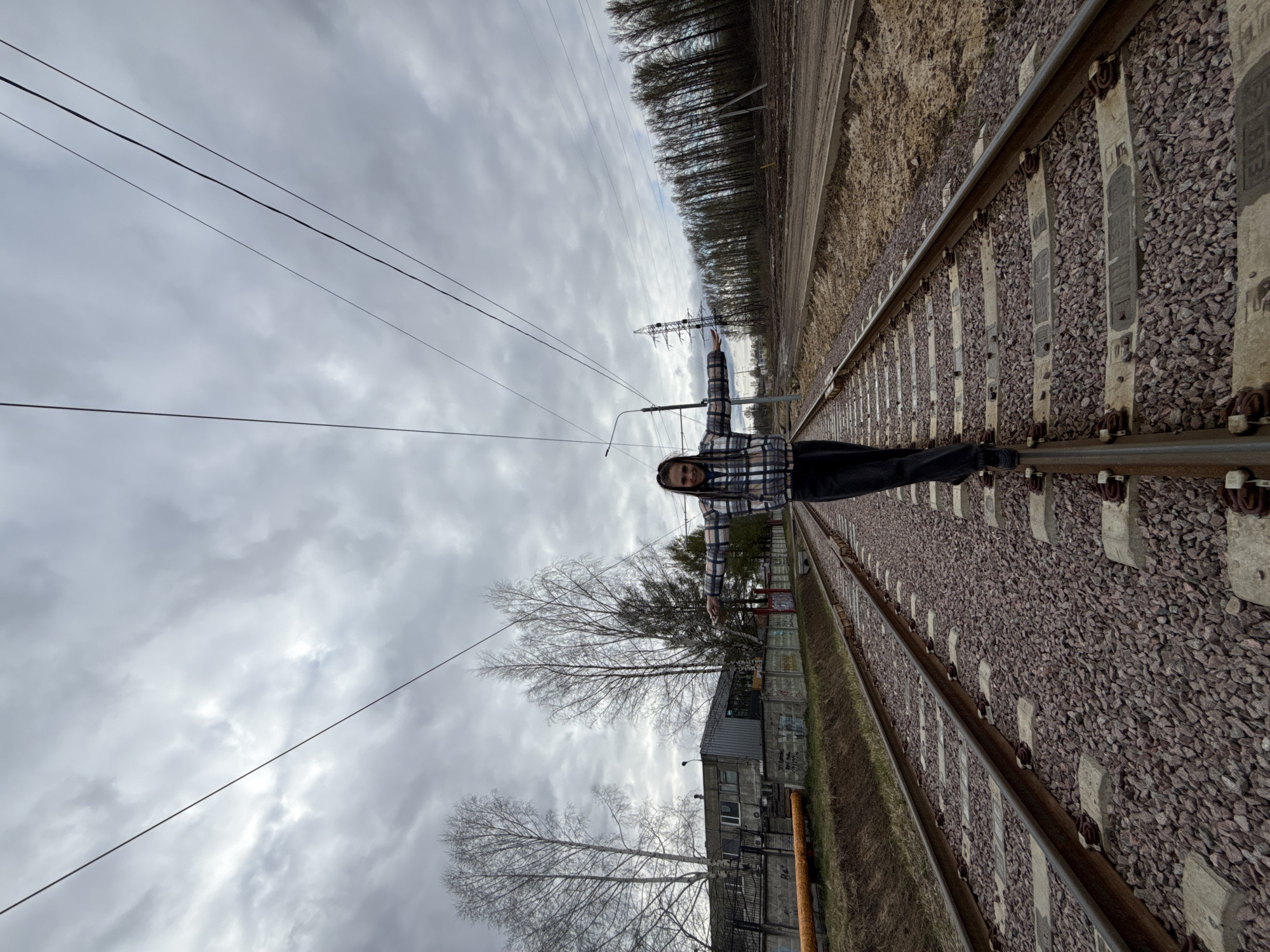 This image, taken in Rusanovo, Russia, is oriented vertically and depicts a person balancing on railway tracks under a cloudy sky.
A young person, wearing a plaid shirt and dark pants, stands with arms outstretched on one of the metal rails of the railway track. They are looking towards the camera with a neutral expression. The railway tracks, constructed with metal rails, concrete sleepers, and grey gravel ballast, stretch diagonally from the foreground into the distance. Faint embossed text, including numbers like "1988" and letters such as "BC" or "BE", is visible on the concrete sleepers.
Parallel to the tracks, to the right of the person, is a strip of brown earth and a dense line of tall, bare deciduous trees, suggesting a forest edge. A large metal power pylon is visible beyond these trees in the middle ground. To the left of the tracks and closer to the foreground, a low-rise, grey building with a dark roof and possibly some graffiti is visible, surrounded by a few bare trees. Multiple power lines traverse the grey, overcast sky.
The weather is cloudy, and the barren state of the trees indicates the season is likely late autumn, winter, or early spring. The overall scene portrays an outdoor, possibly semi-industrial or rural setting adjacent to an active railway line.