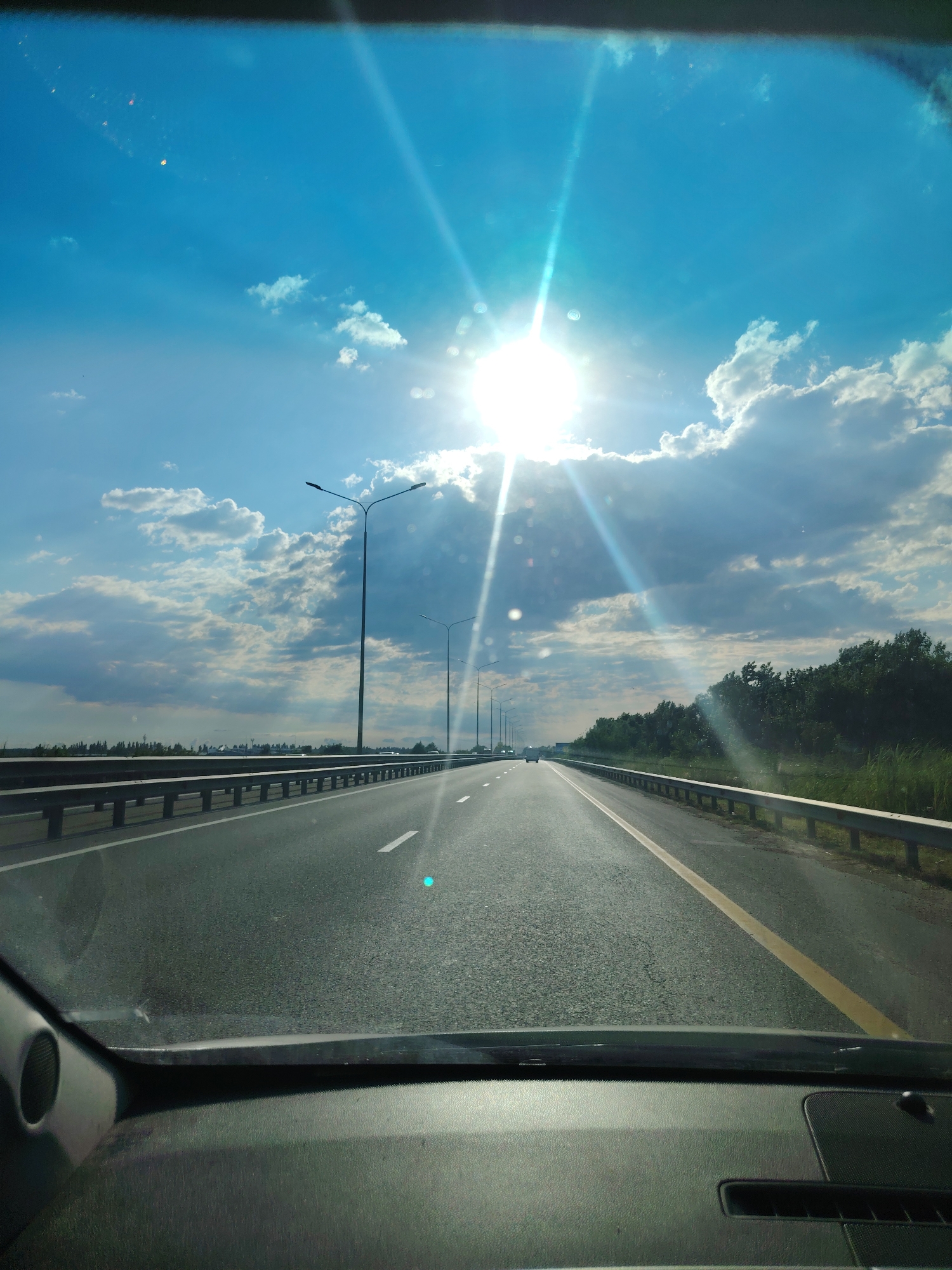 The image captures a view from inside a vehicle looking forward onto a highway in the city of Prikubanskiy, Russia. The sun is bright and high in a clear blue sky, with scattered white clouds. Sunbeams radiate outwards, creating a lens flare effect. The highway has multiple lanes, separated by white lines, and a yellow line on the right side. Guardrails run along the sides of the road, and streetlights are visible at intervals. To the right of the highway, there is a dense line of trees. In the distance, there are faint signs of urban infrastructure and some vehicles on the road. The interior of the vehicle, including the dashboard and a speaker, is visible in the foreground, obscuring the bottom portion of the scene.
