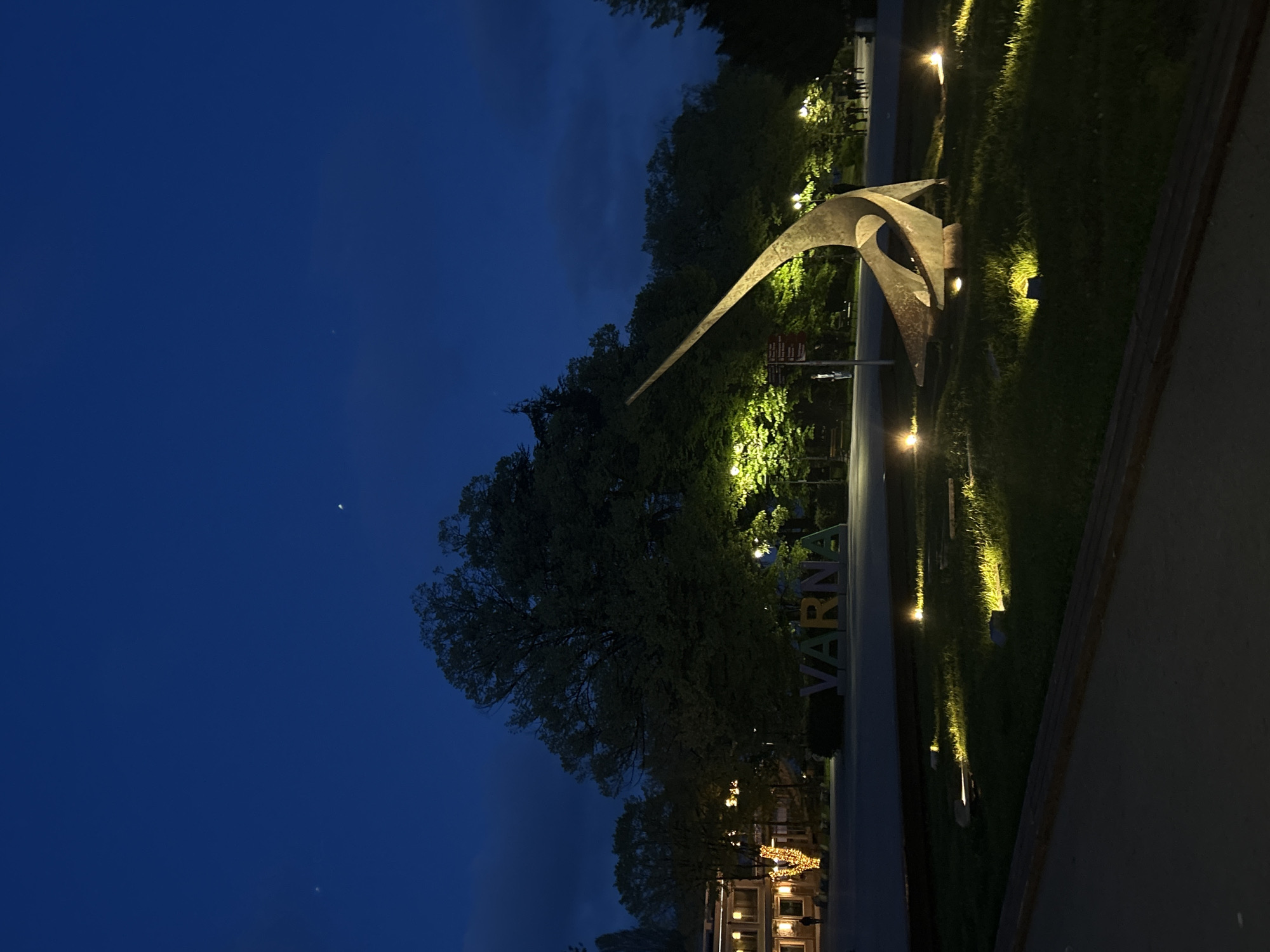 The image depicts a scene in Varna, Bulgaria, at night. The sky is a deep blue, with a faint crescent moon visible. In the foreground, a modern metal sculpture stands illuminated by spotlights. Behind the sculpture, the word "VARNA" is spelled out in large, colorful letters. The trees and grass surrounding the sculpture are also lit by spotlights. In the background, there are buildings with lights on, suggesting a lively urban environment. No people are visible.