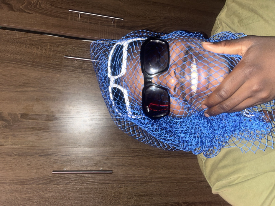 A person with dark skin lies down with their head tilted to the right. A blue fishnet covers their face and neck. Two pairs of sunglasses, one with a white frame and one with a black frame, rest on their forehead and nose, respectively. Their hand holds the net in place, partially obscuring their face. A dark wood-grain surface with drawer pulls is visible in the background. The person wears light green or khaki-colored clothing.