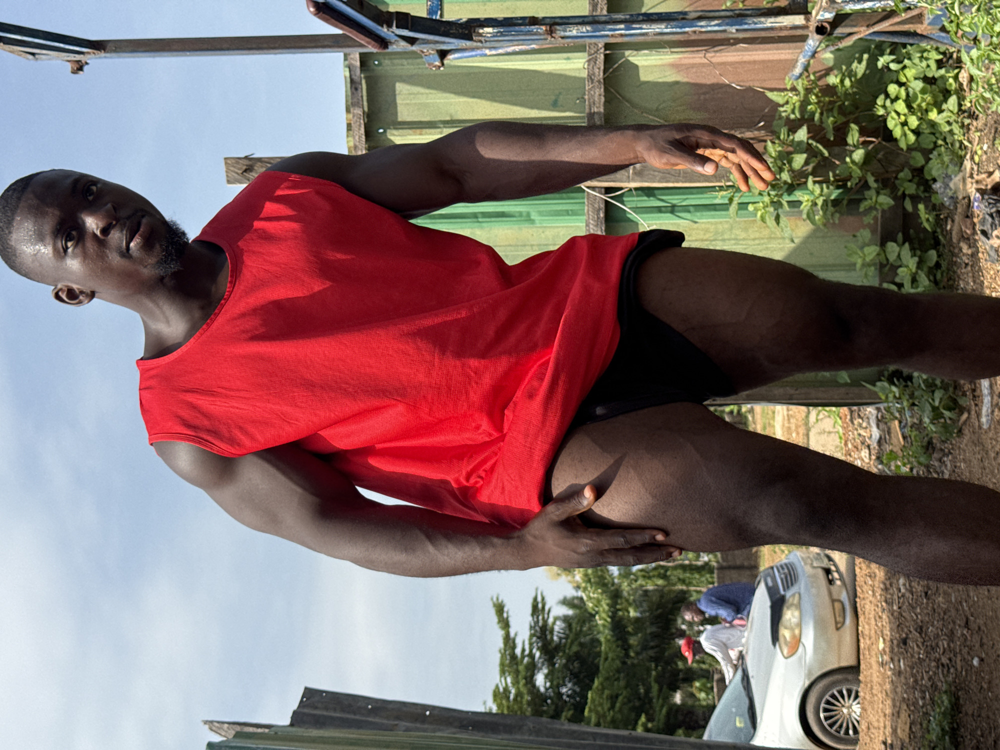 A Black man with muscular legs and arms is standing outdoors, looking down and to his right, with a slight smirk on his face. He is wearing a red sleeveless shirt and black shorts. His right hand is resting on his left thigh, and his left arm is bent at the elbow. Behind him, to his left, is a silver sedan with two men standing near it. One man is wearing a red cap and a white shirt, and the other is wearing a blue shirt. To the right of the man in the foreground, there are green metal walls and some overgrown vegetation. The sky is visible in the background, with some clouds. The overall setting appears to be an outdoor area, possibly a street or a yard, in Akwanga, Nigeria.